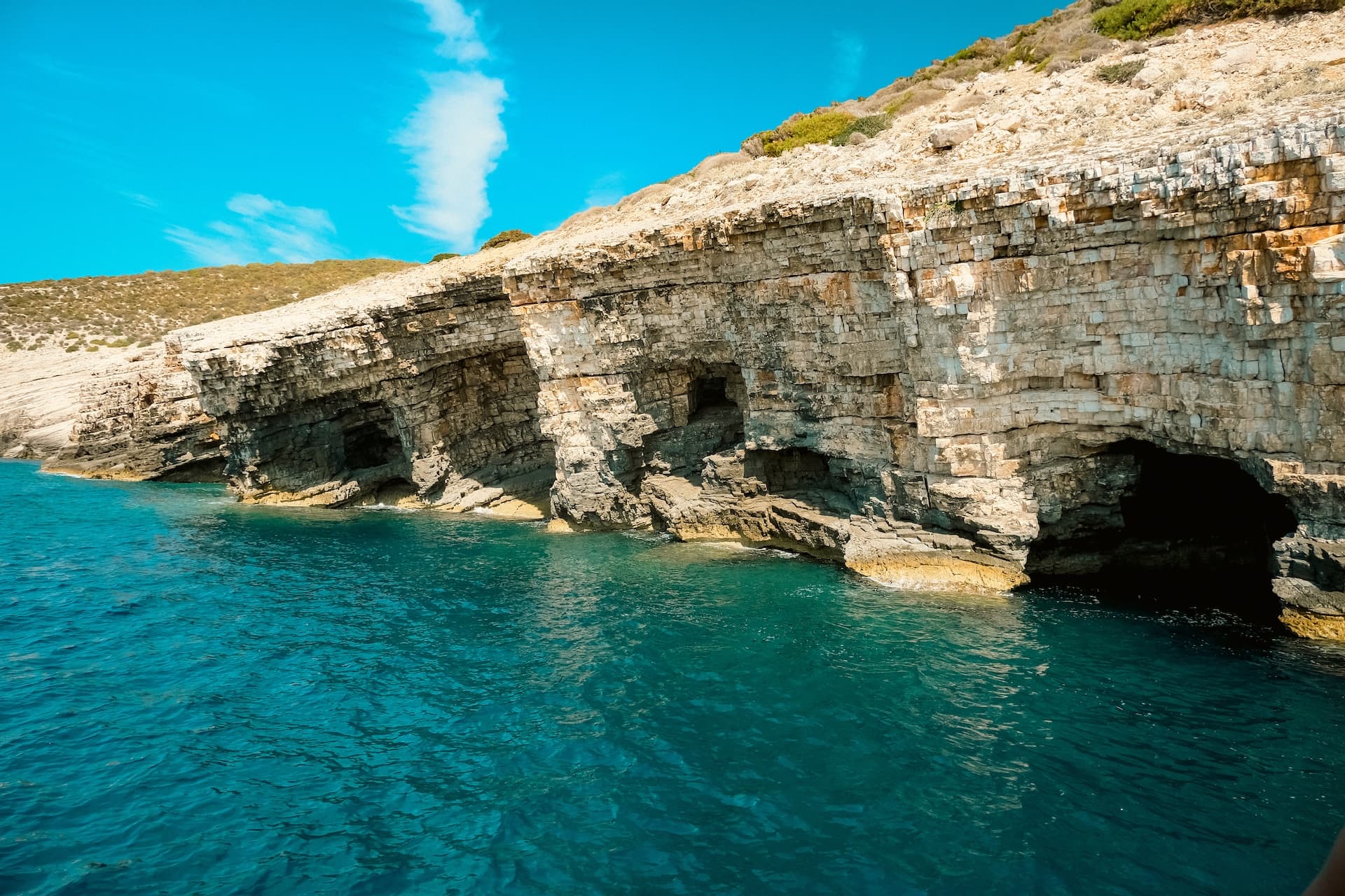 Sea caves in layered limestone cliffs above turquoise water near Komiza, Vis island.