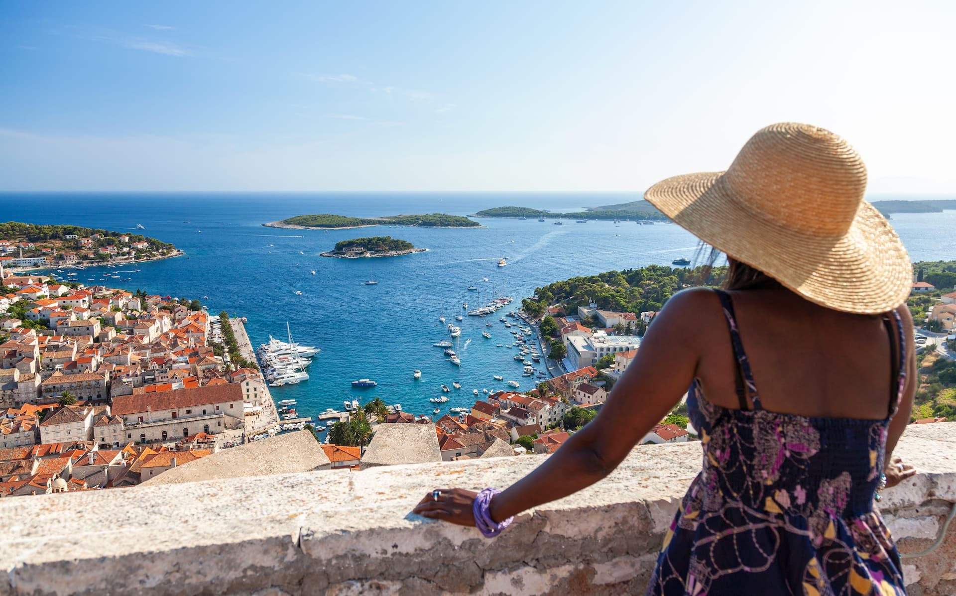 Woman in sun hat overlooks Hvar Island town, harbor, and blue Adriatic Sea.