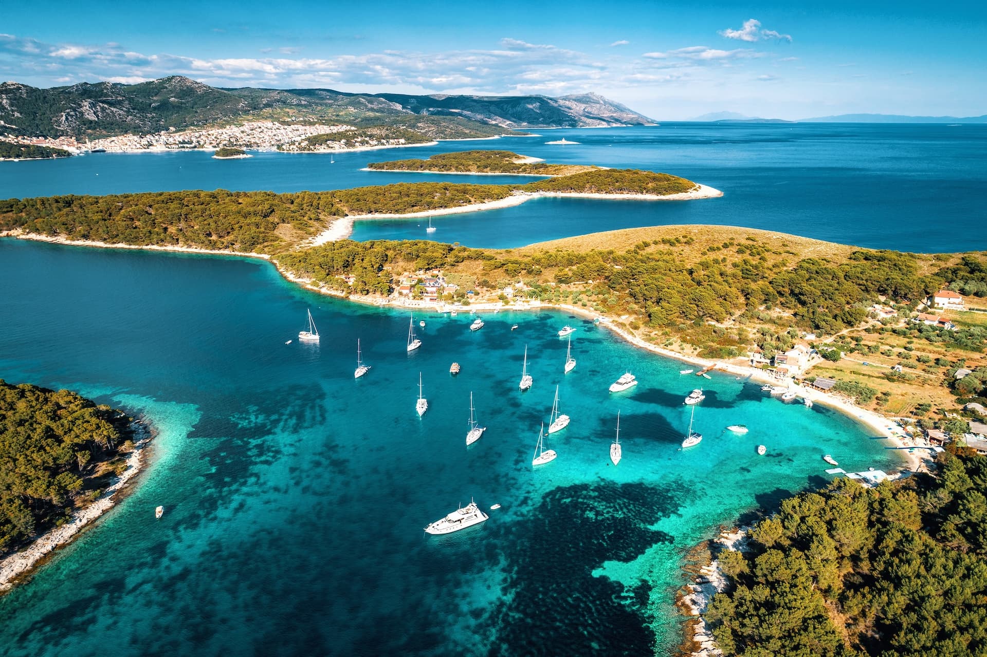 Boats and yachts docking in turquoise cove near Pakleni Islands with forested hills and distant town.