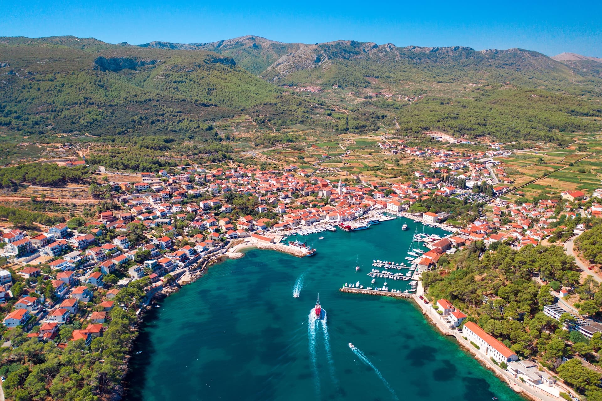 Coastal town with red-roofed buildings nestled below green mountains overlooking a turquoise harbor with boats.