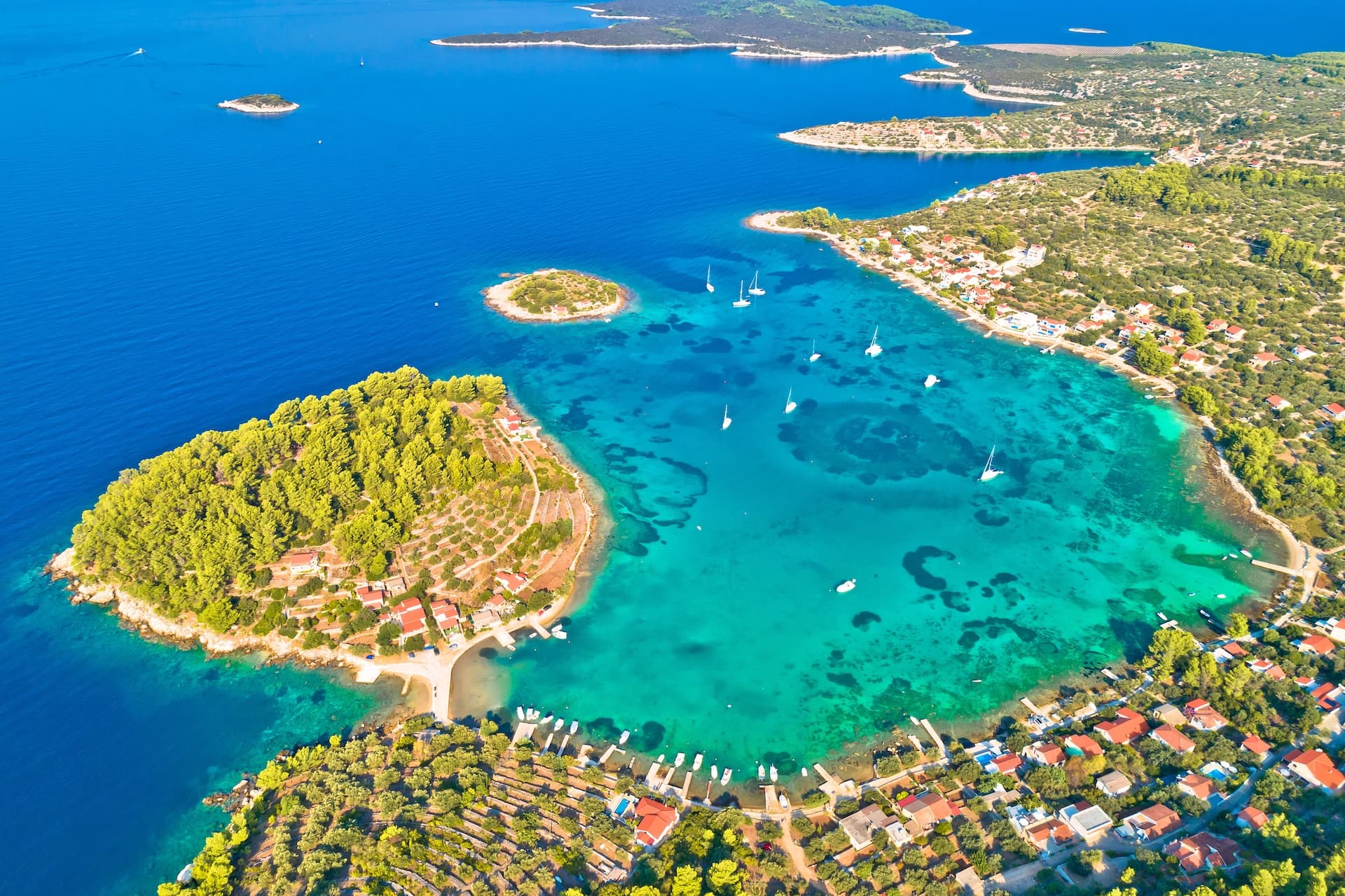 Aerial view of Korčula island bay with turquoise water, sailboats, and coastal village homes.