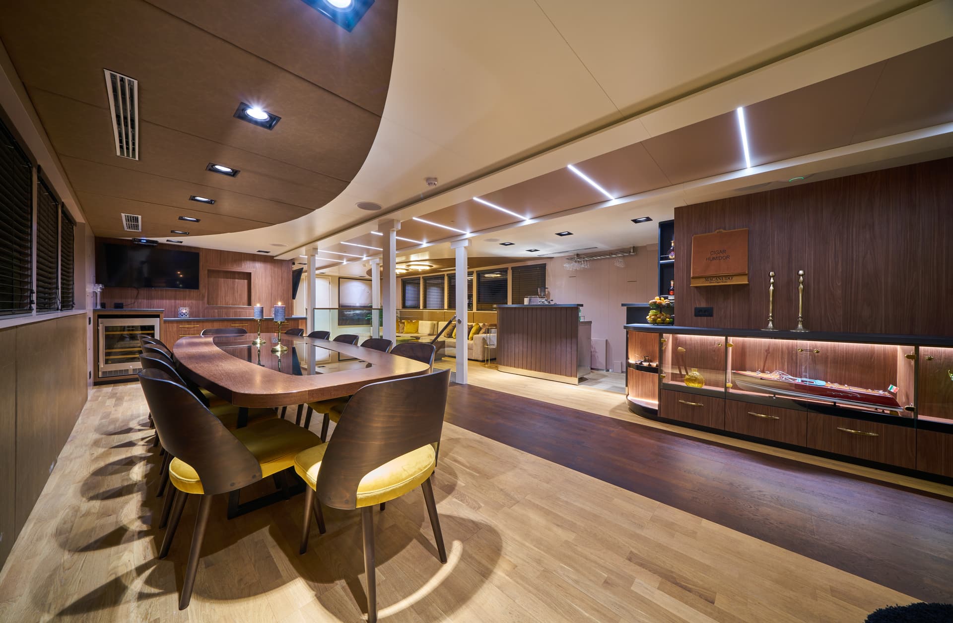 Luxury yacht saloon with curved dining table, wood paneling, and illuminated display case with model boat.