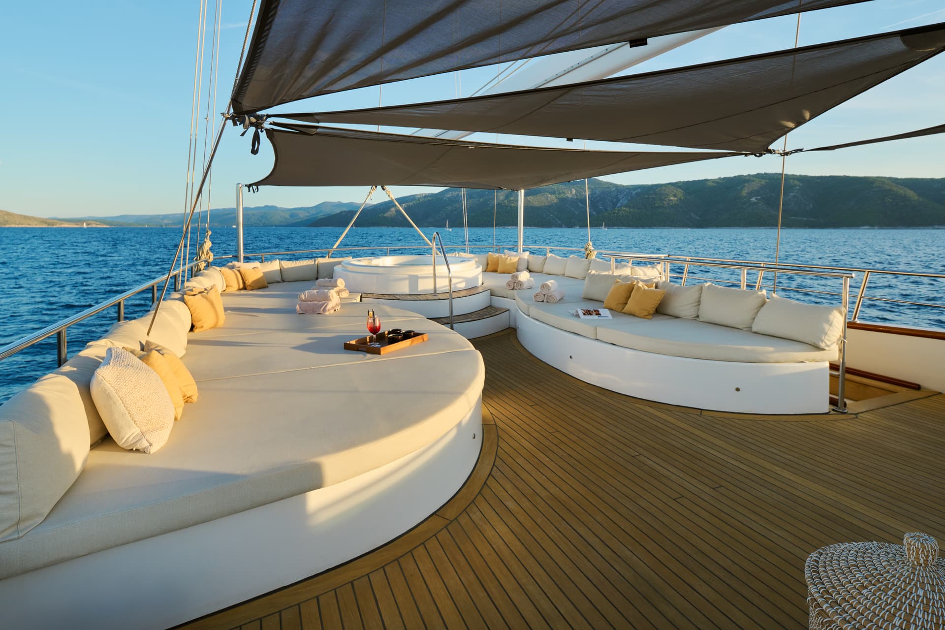 Yacht sundeck with lounge seating, hot tub, and cocktails overlooking green mountains and blue sea.