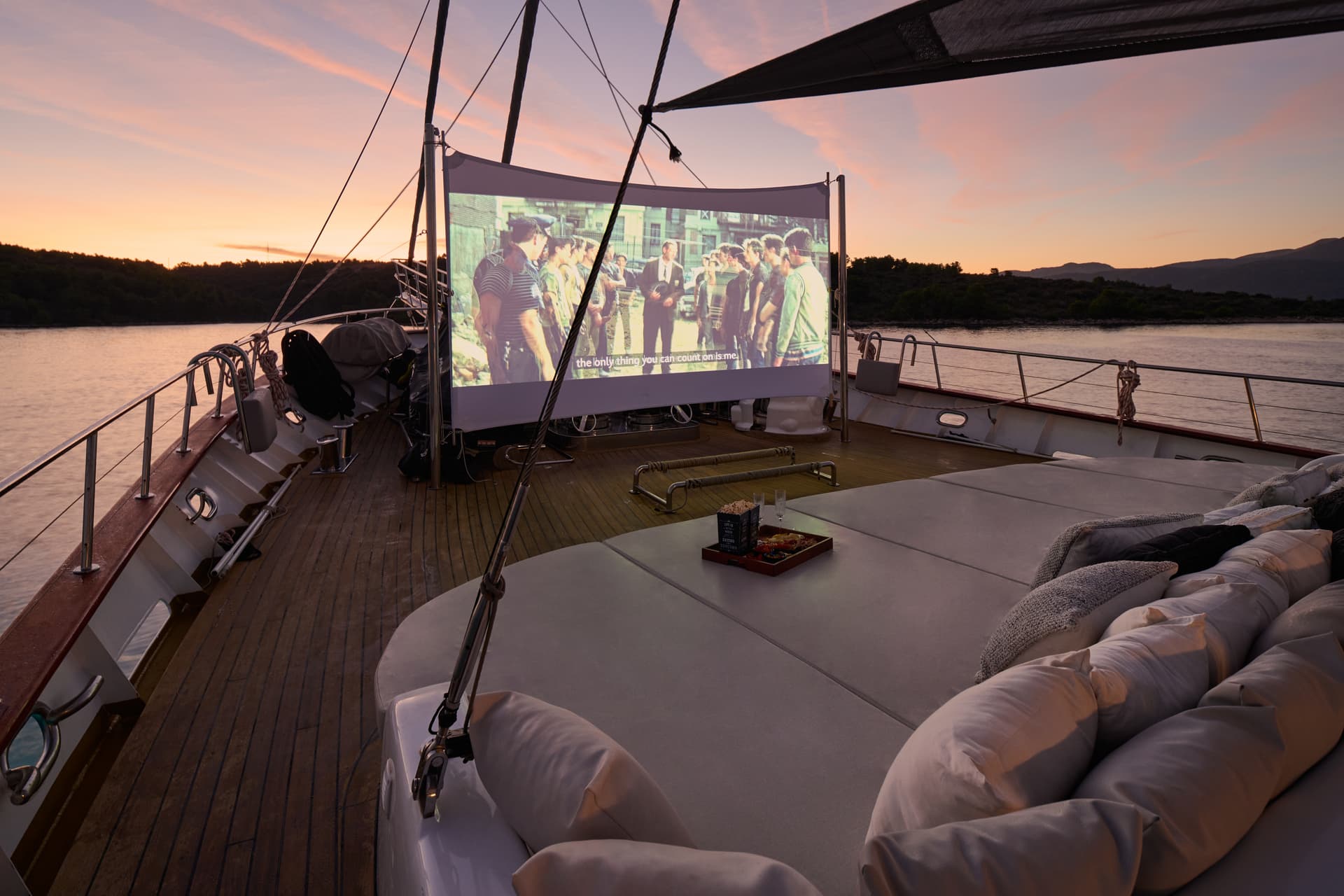 Outdoor movie night on a yacht deck with sunset over mountains and water.