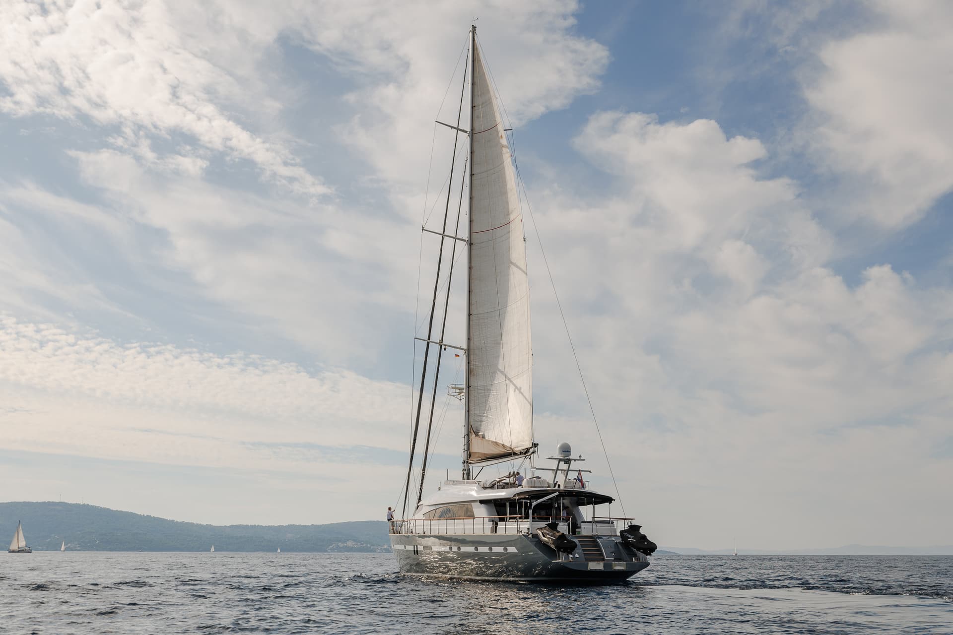 Large sailboat with sails up on choppy water, stern view, with green coastline in background.