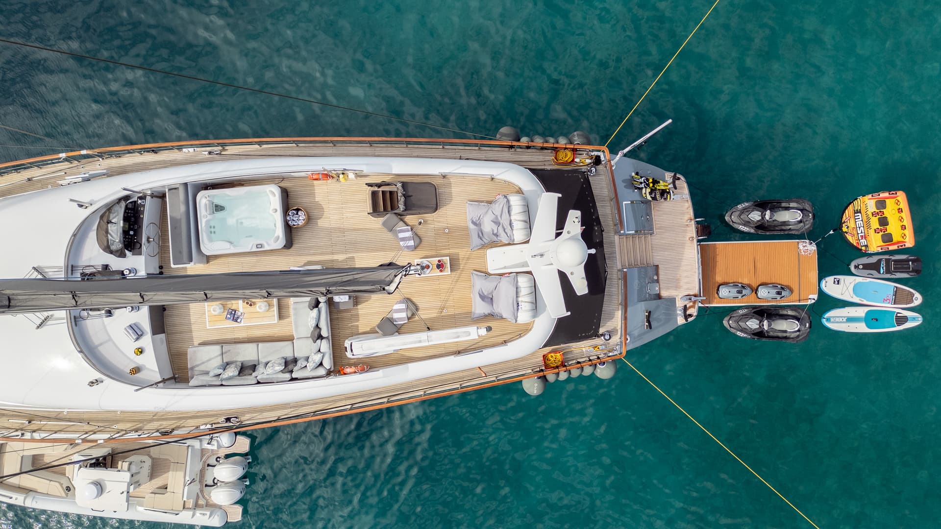 Aerial view of a large yacht deck with a hot tub, lounge area, and water toys in clear turquoise water.