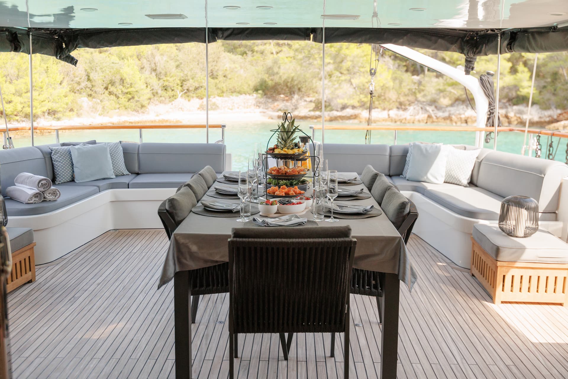 Outdoor dining setup on yacht deck with fruit platter, set table, and green coastline view.