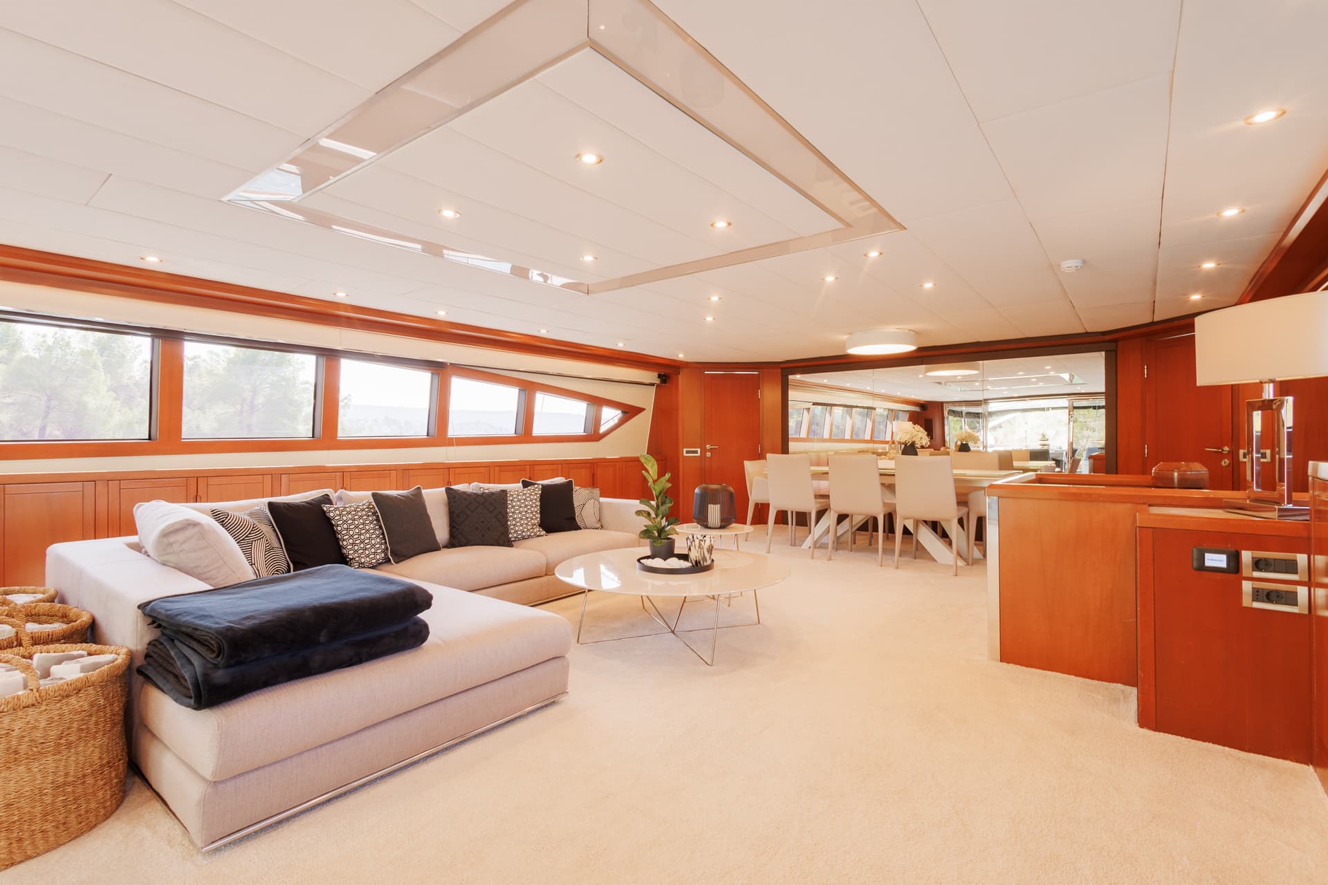 Luxury yacht interior with light sofa, dining area, wood paneling, and view of trees.