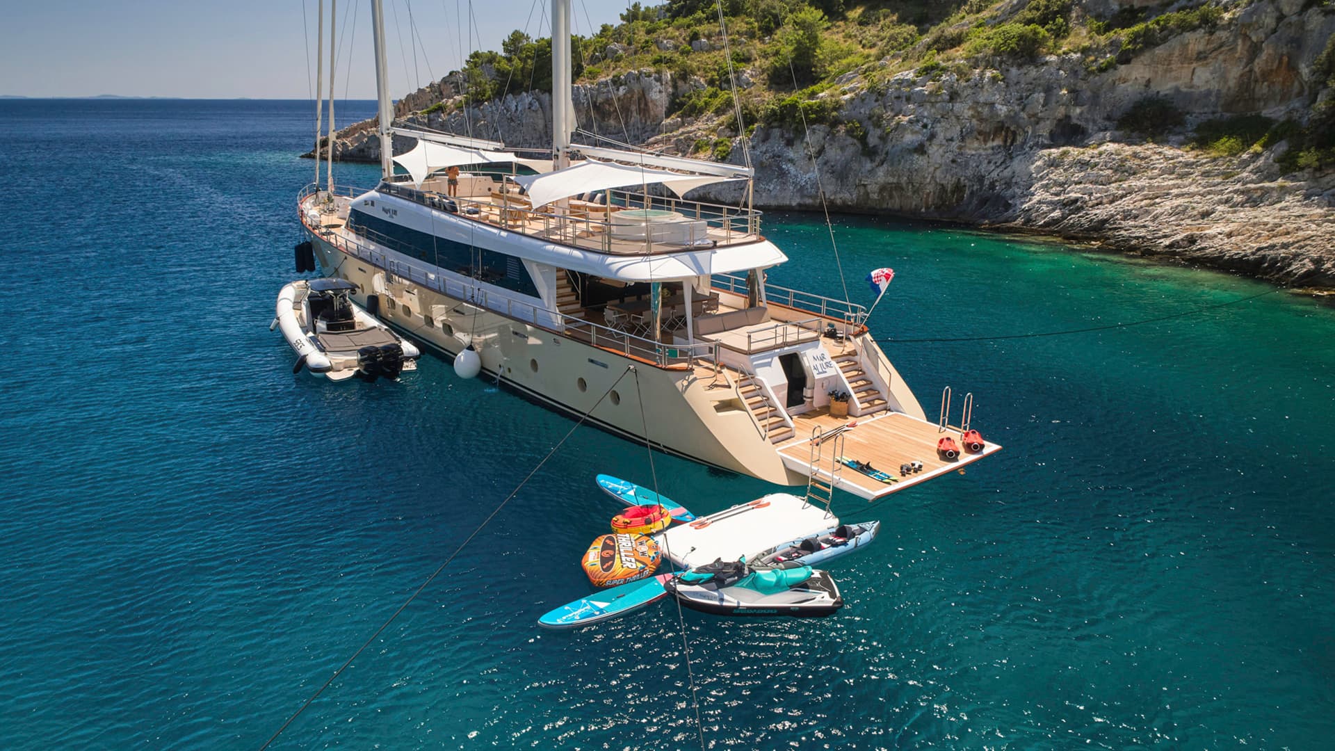 Large yacht anchored near rocky coastline with clear turquoise water and water toys.