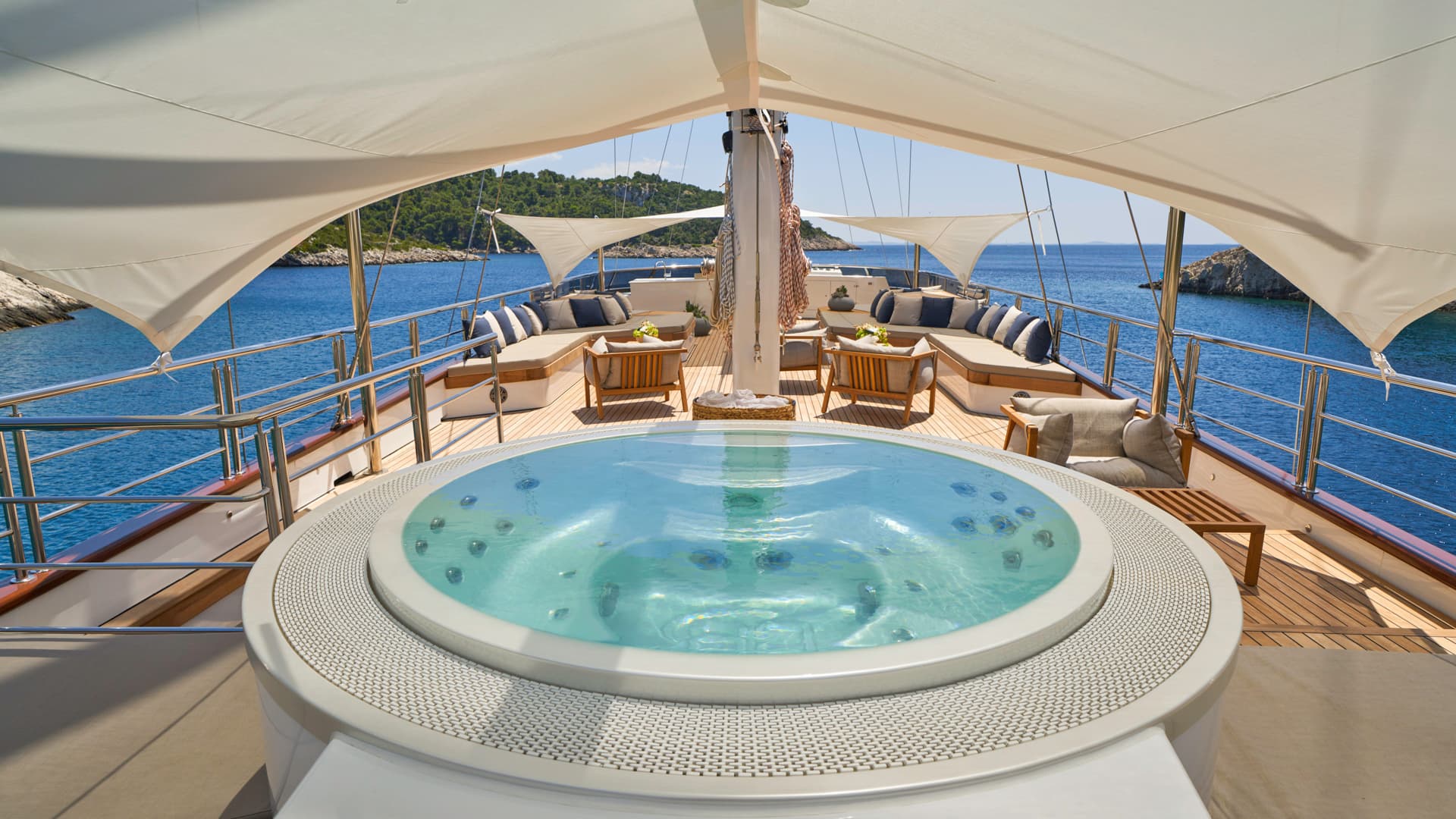 Jacuzzi on yacht deck with lounge seating overlooking blue sea and green coastline