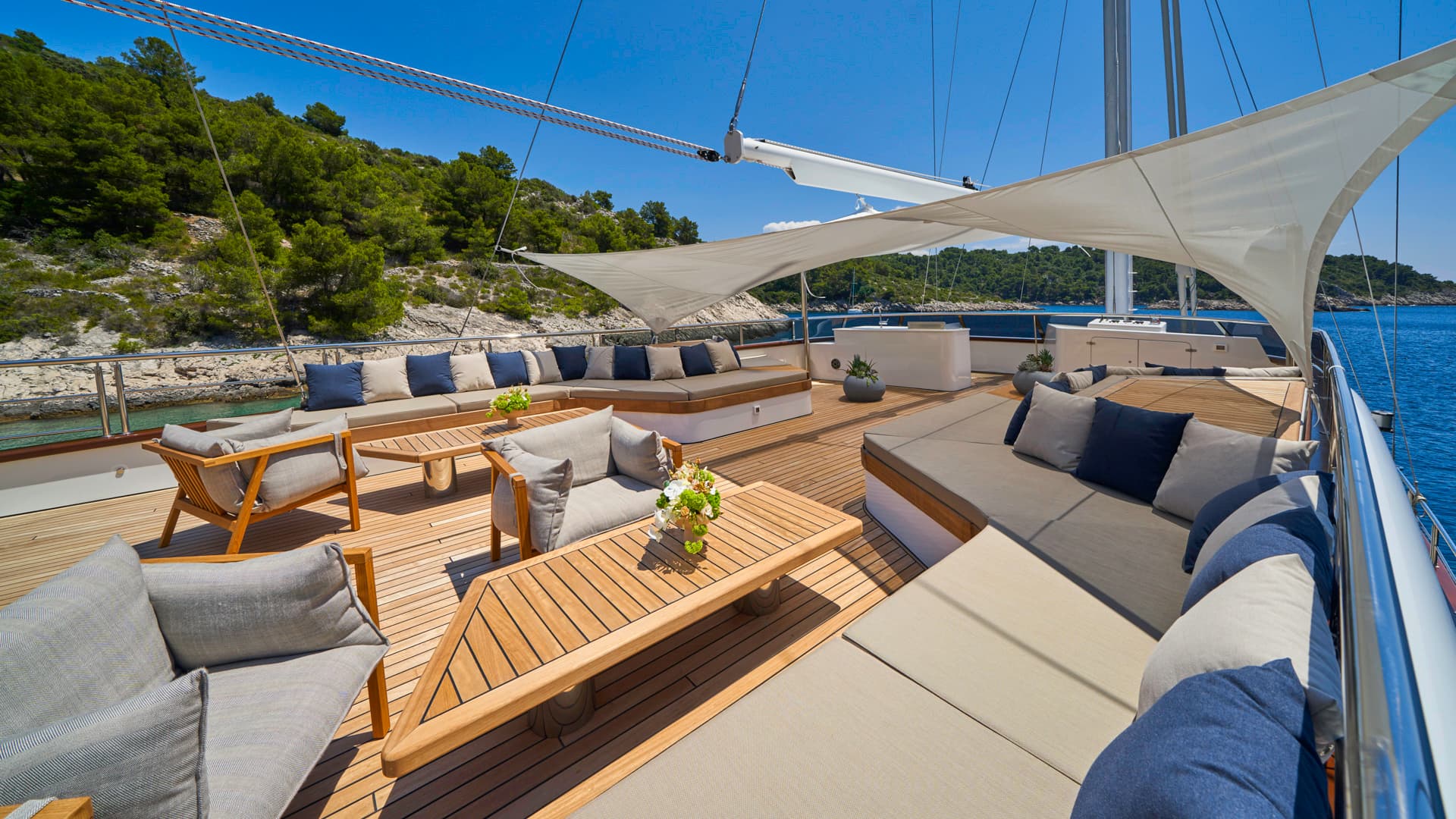 Luxury yacht sun deck with teak flooring, shaded lounge seating, and green coastline nearby.