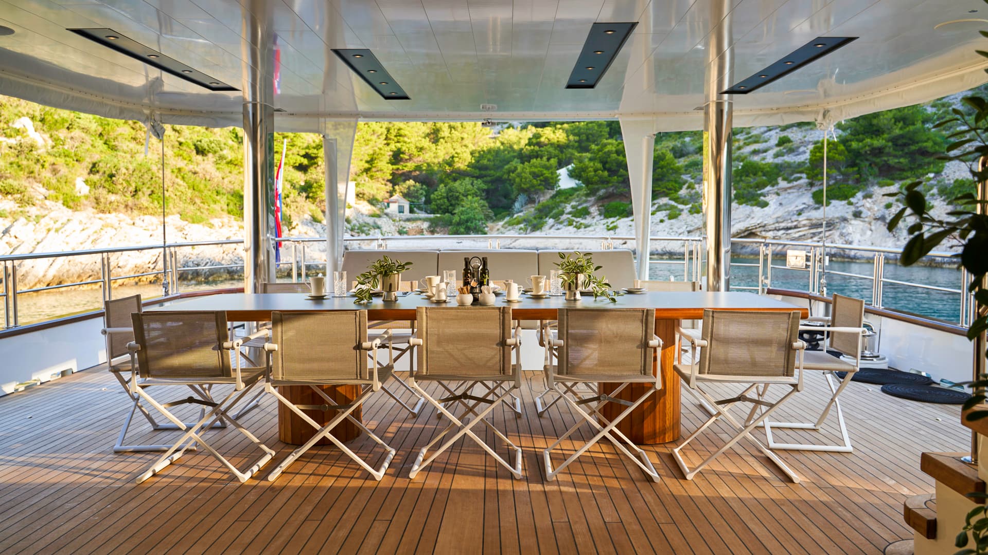 Outdoor dining table set on a yacht deck overlooking a rocky, green coastline.