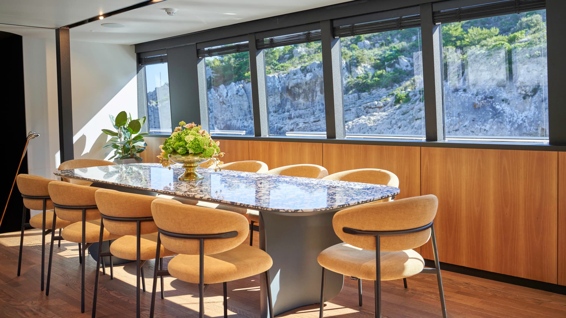 Dining table with marble top and tan chairs inside, viewing rocky, green hillside through windows.