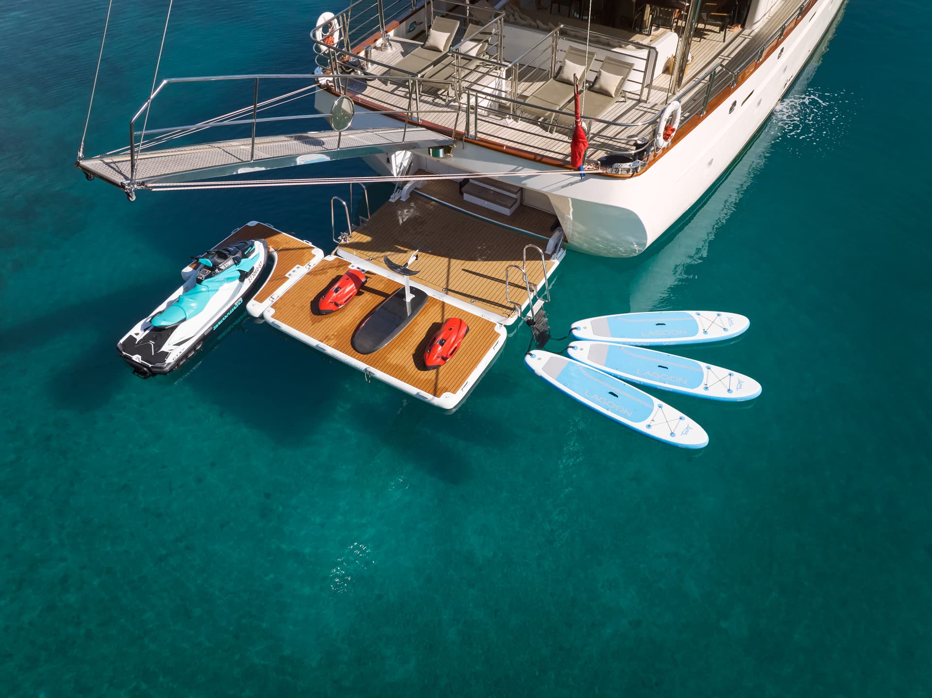 Yacht stern with jet skis, sea scooters, and paddleboards floating in clear turquoise water.