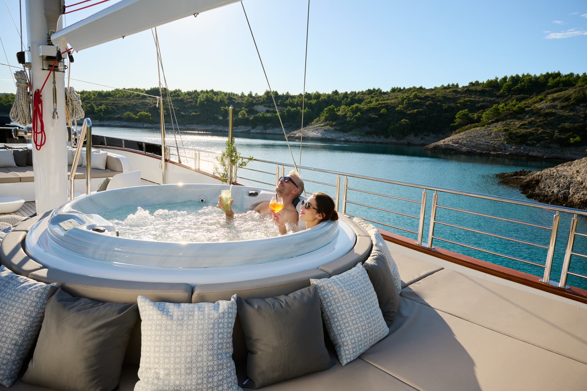 Couple relaxing in yacht jacuzzi with drinks near tree-covered coastline.