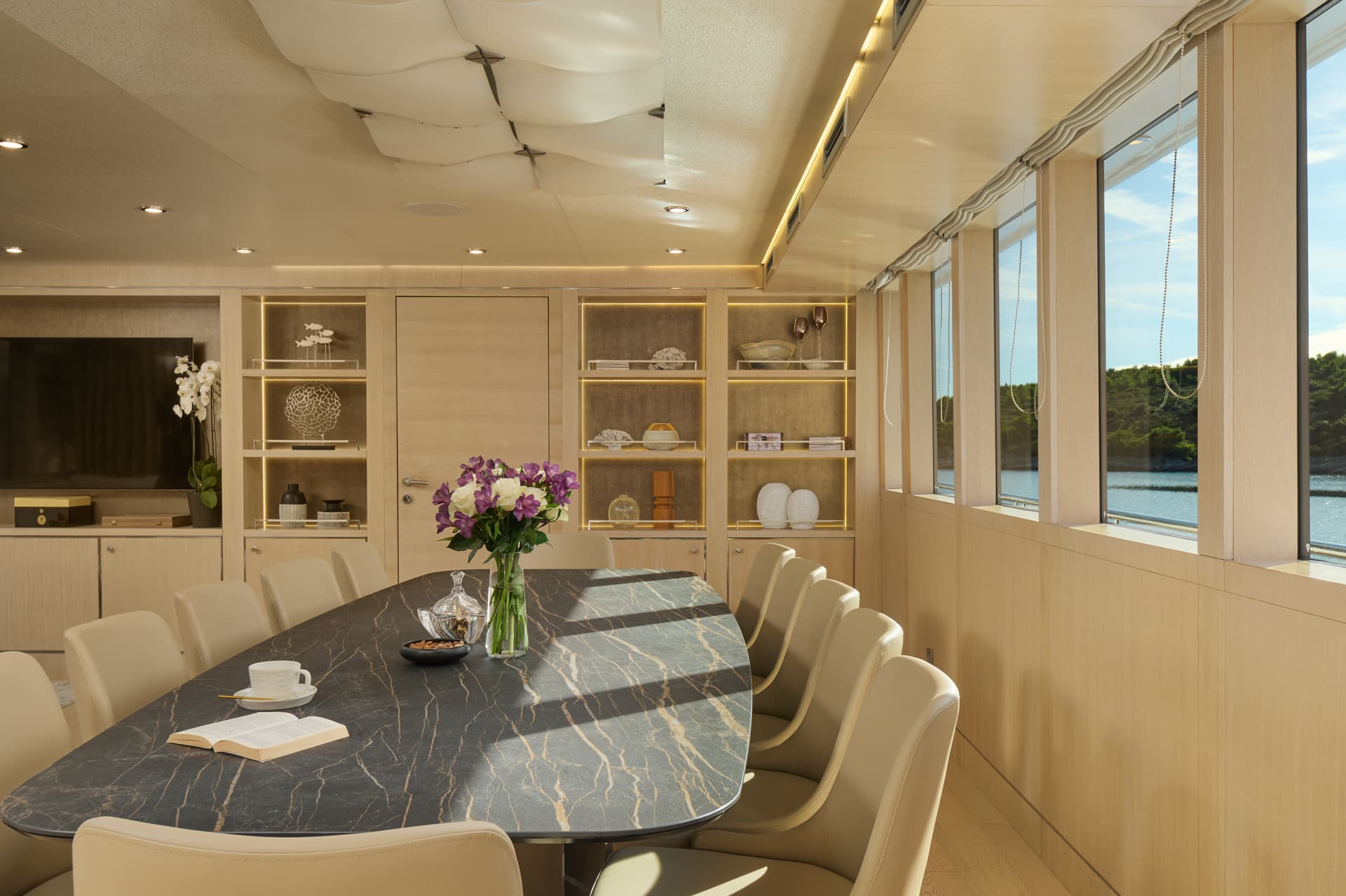 Dining area on a yacht with marble table, flowers, and view of green coastline and water.