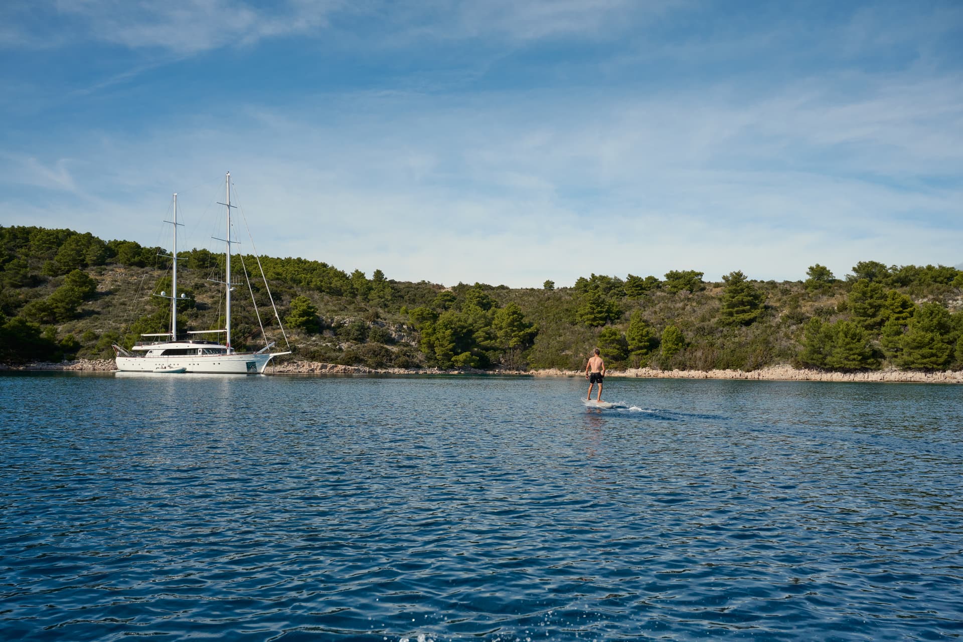 Man wakesurf boarding near a large white sailboat anchored by a wooded coastline.
