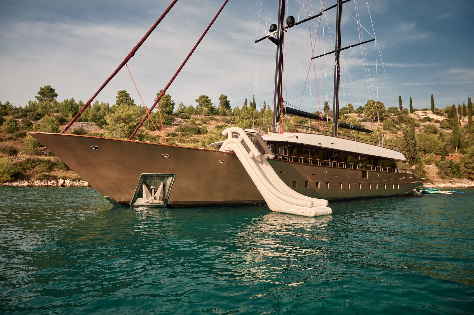 Large yacht with inflatable waterslide anchored near a lush, rocky coastline.