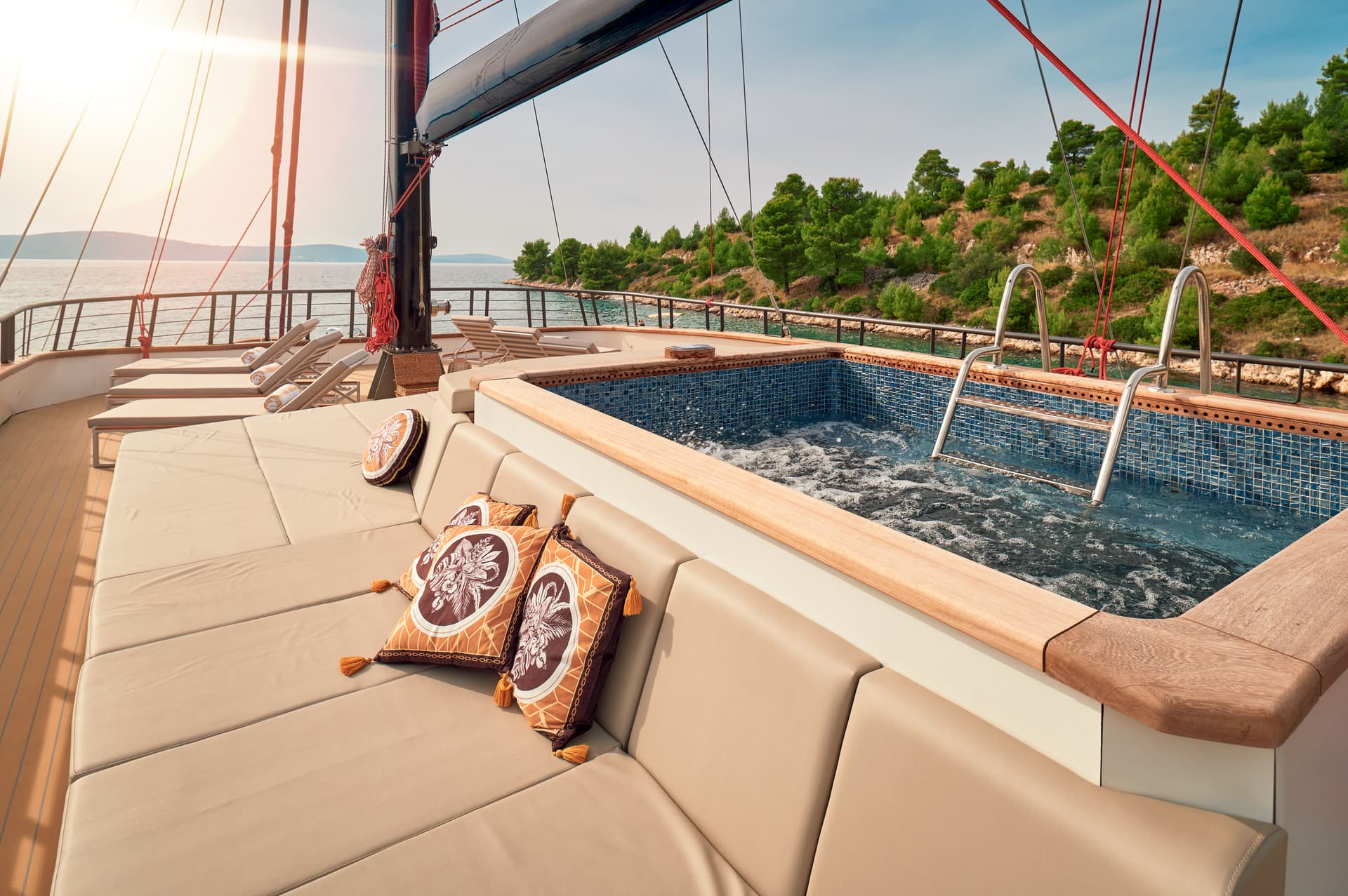 Yacht sundeck with jacuzzi, sun loungers, and pillows near a tree-covered coastline.