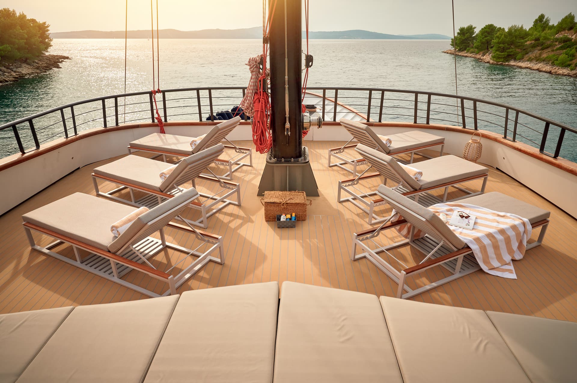 Sundeck lounge chairs on a yacht sailing past a tree-lined rocky coastline at sunset.
