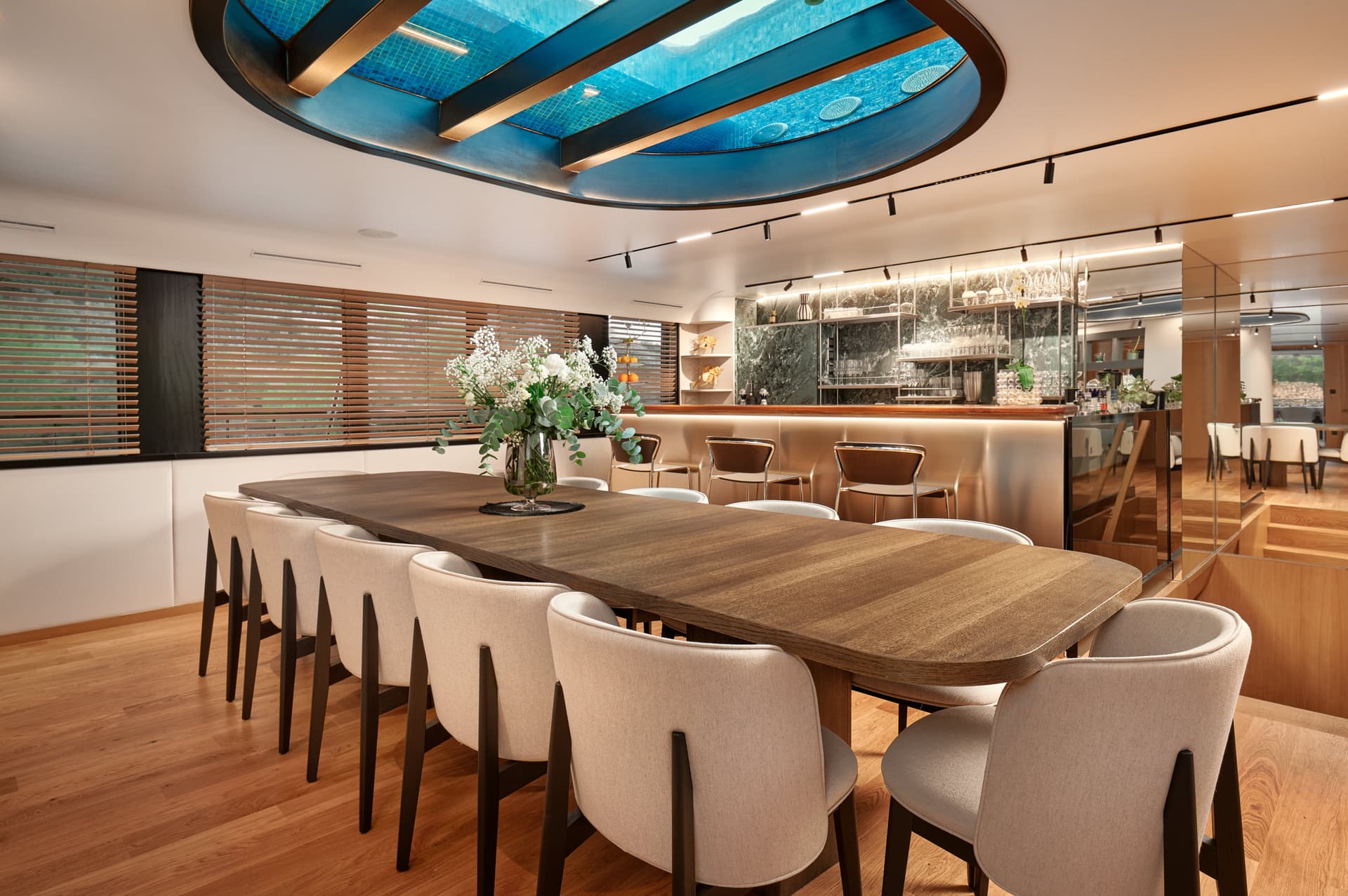 Dining room on a yacht with wood table, beige chairs, and overhead skylight showing blue water.