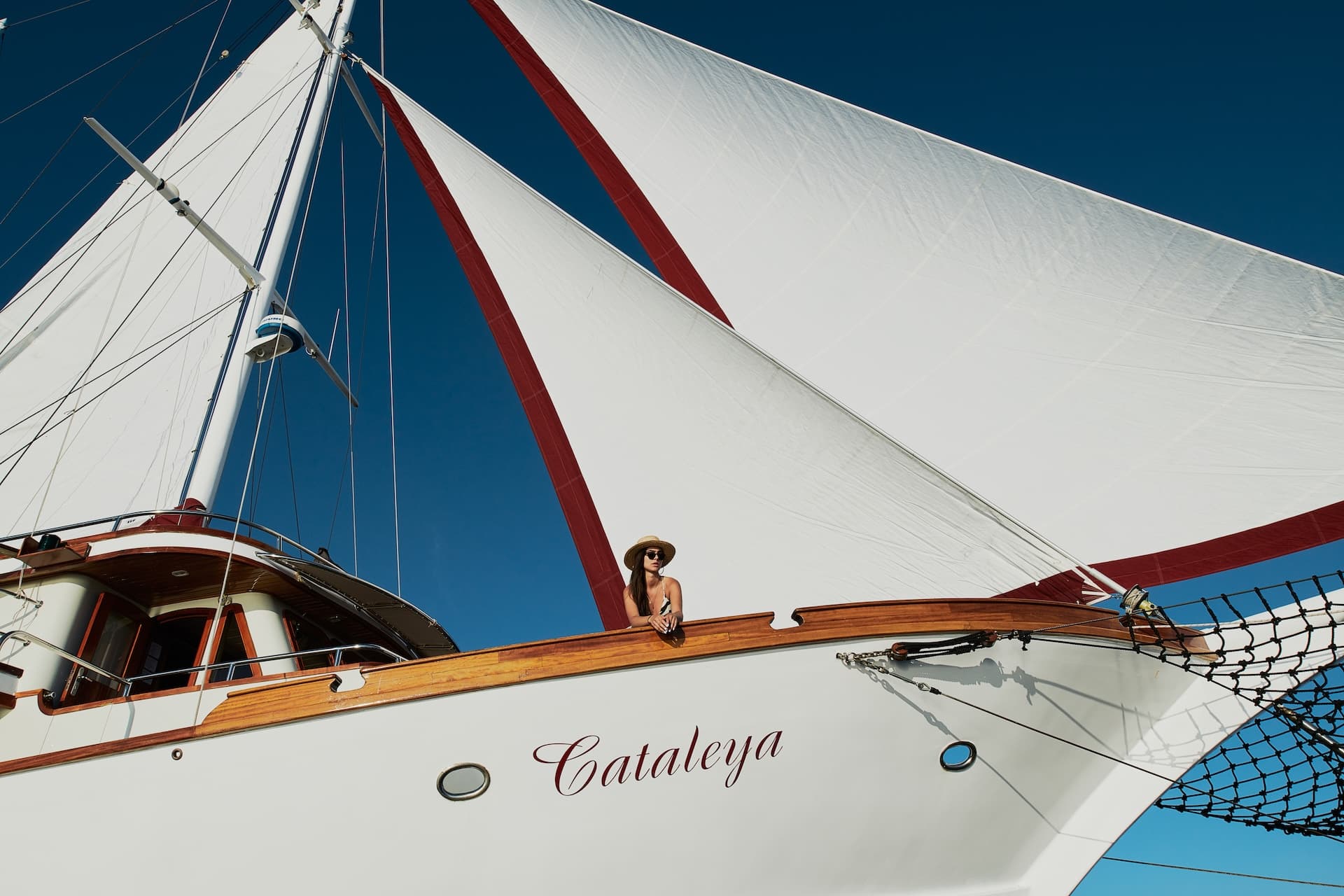 Woman on sailboat named Cataleya with white and red sails against clear blue sky.