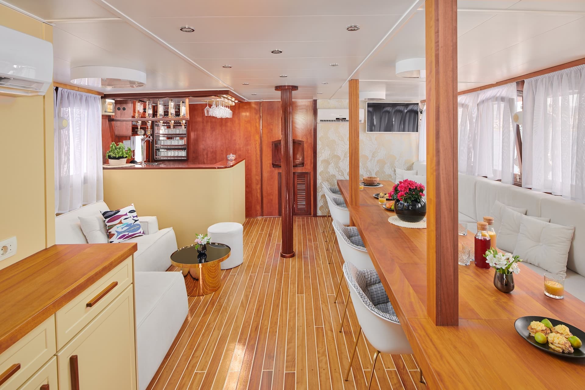 Interior saloon lounge and dining area with wood floors, white seating, and a bar on a boat.
