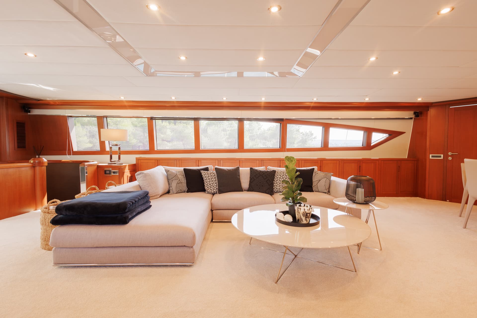 Modern yacht interior salon with beige sectional sofa, wood paneling, and bright windows.