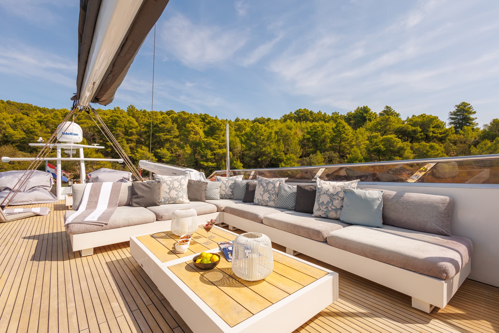 Yacht lounge area with teak deck, white seating, and wooded coastline backdrop.