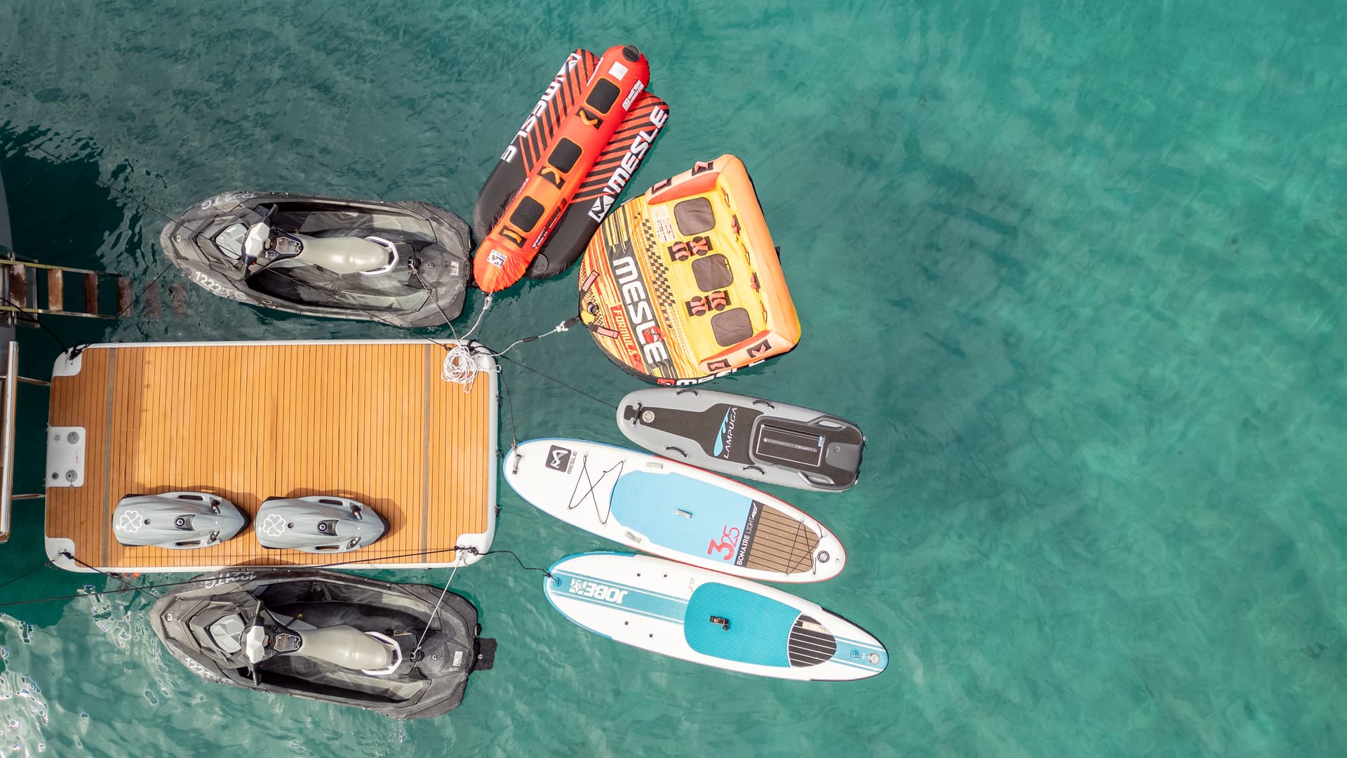 Water toys, jet skis, and paddleboards tied to a wooden dock on clear turquoise water.