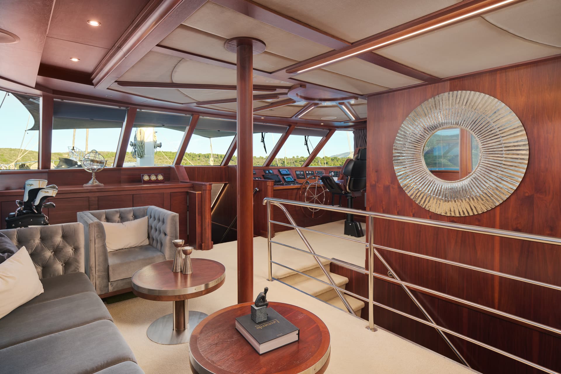Yacht wheelhouse lounge with wood paneling, gray seating, and view of green coastline.