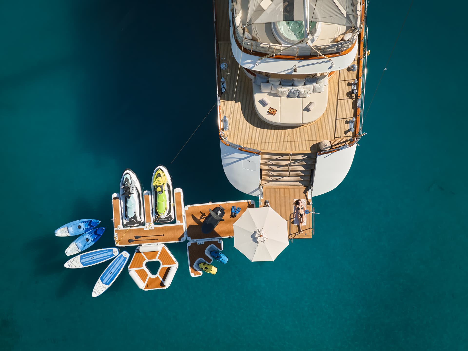 Yacht anchored in turquoise water with inflatable dock, jet skis, kayaks, and woman sunbathing.