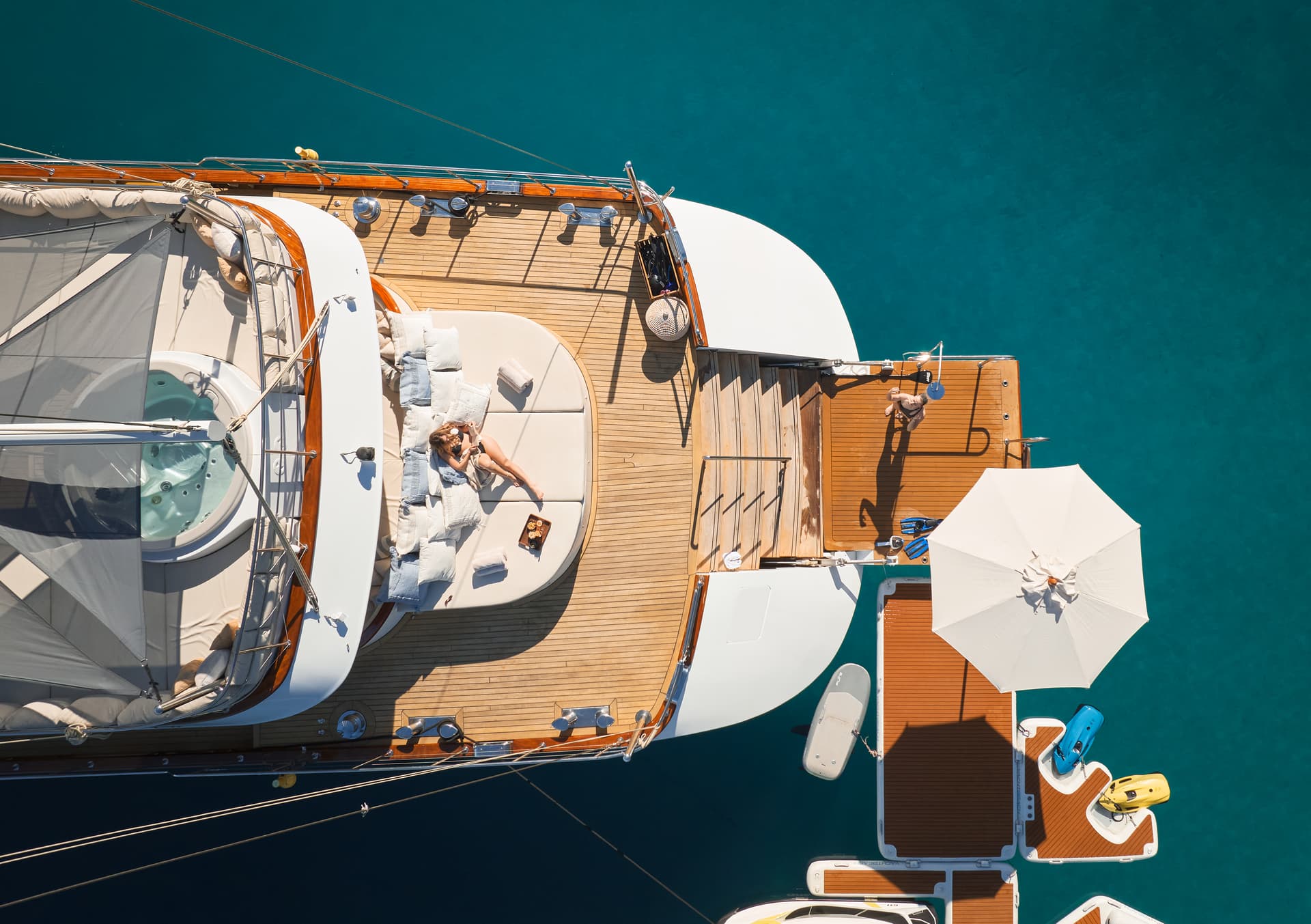 Aerial view of woman relaxing on yacht sunpad next to hot tub over turquoise water with swim platform.