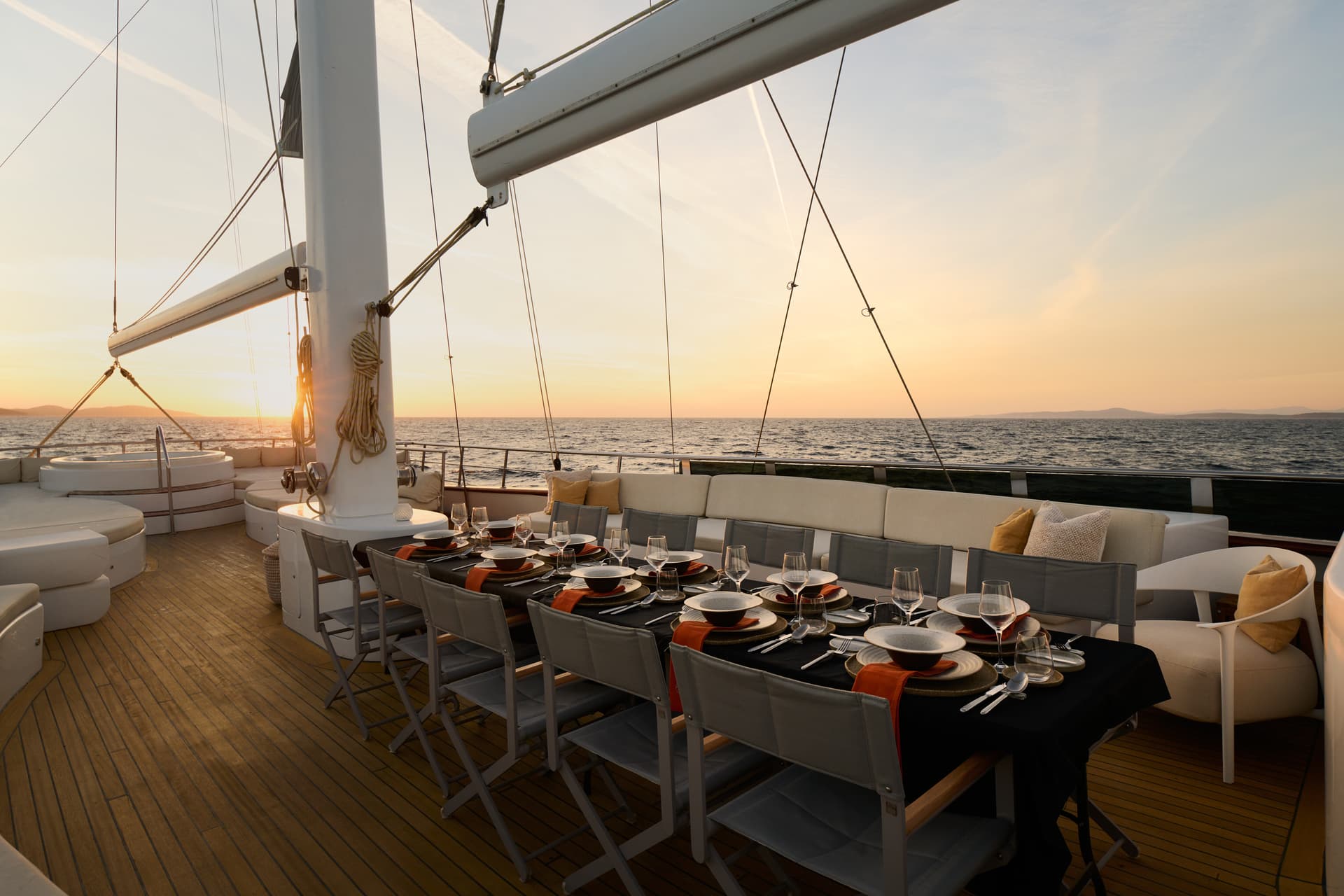 Outdoor dining setup on a yacht deck at sunset with distant mountains over the sea.