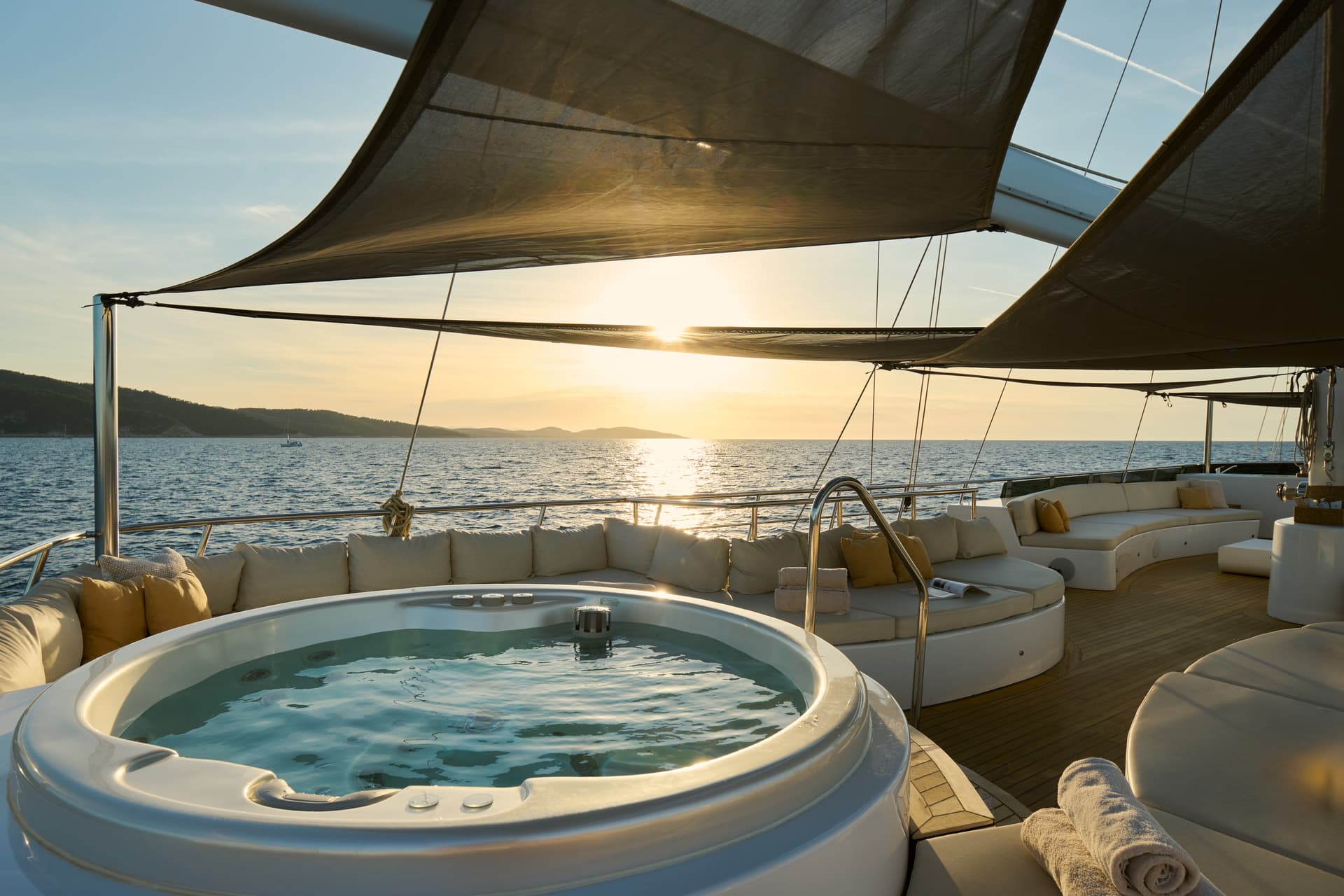 Jacuzzi and lounge seating on a yacht deck overlooking the sea at sunset with hilly coastline.