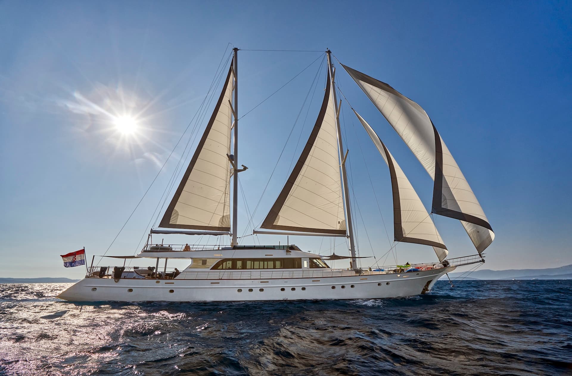White motor sailer with sails up on dark blue, sunlit sea near distant coastline.