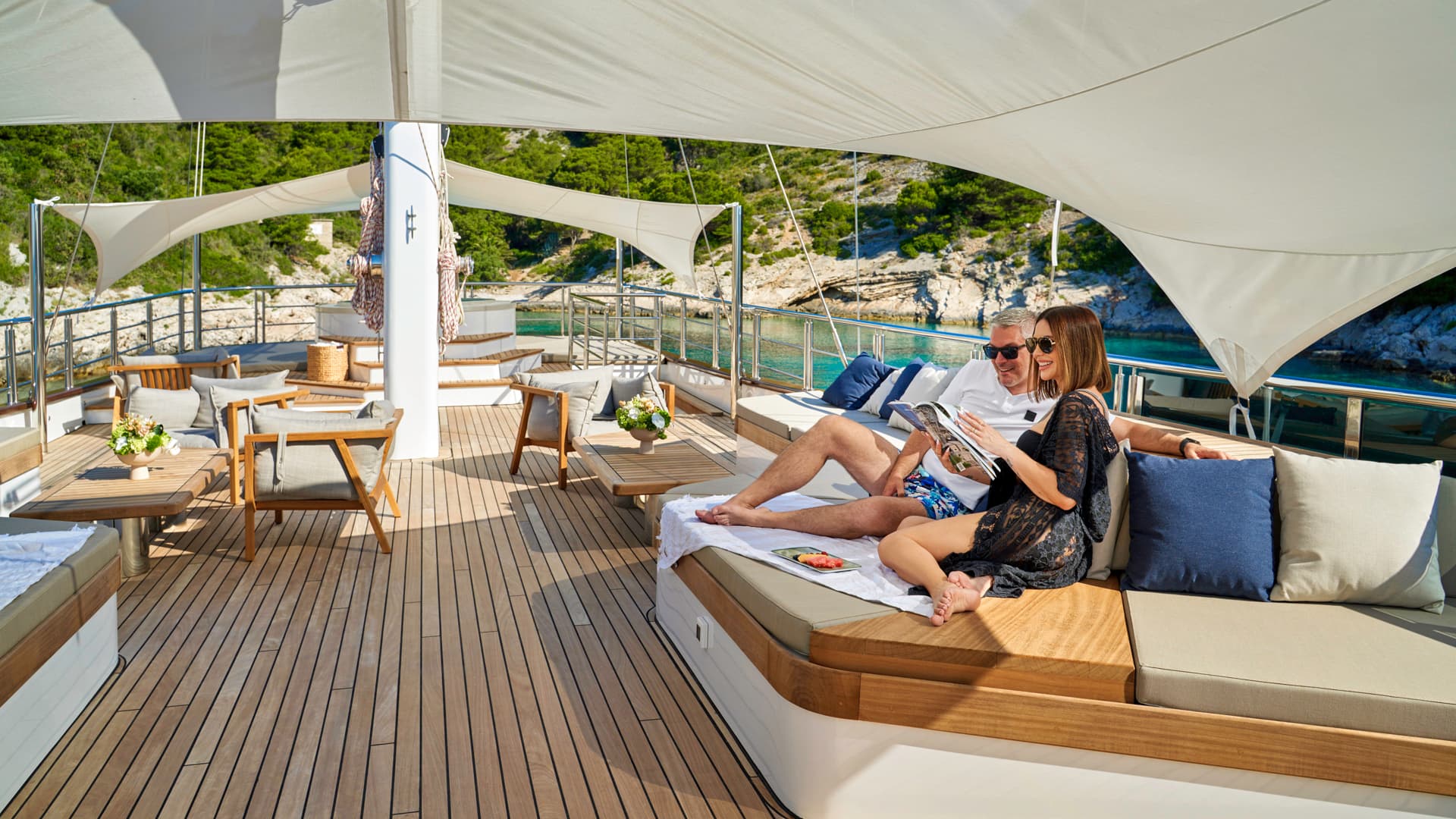 Couple relaxing on yacht deck with wooden flooring near a lush, rocky coastline.