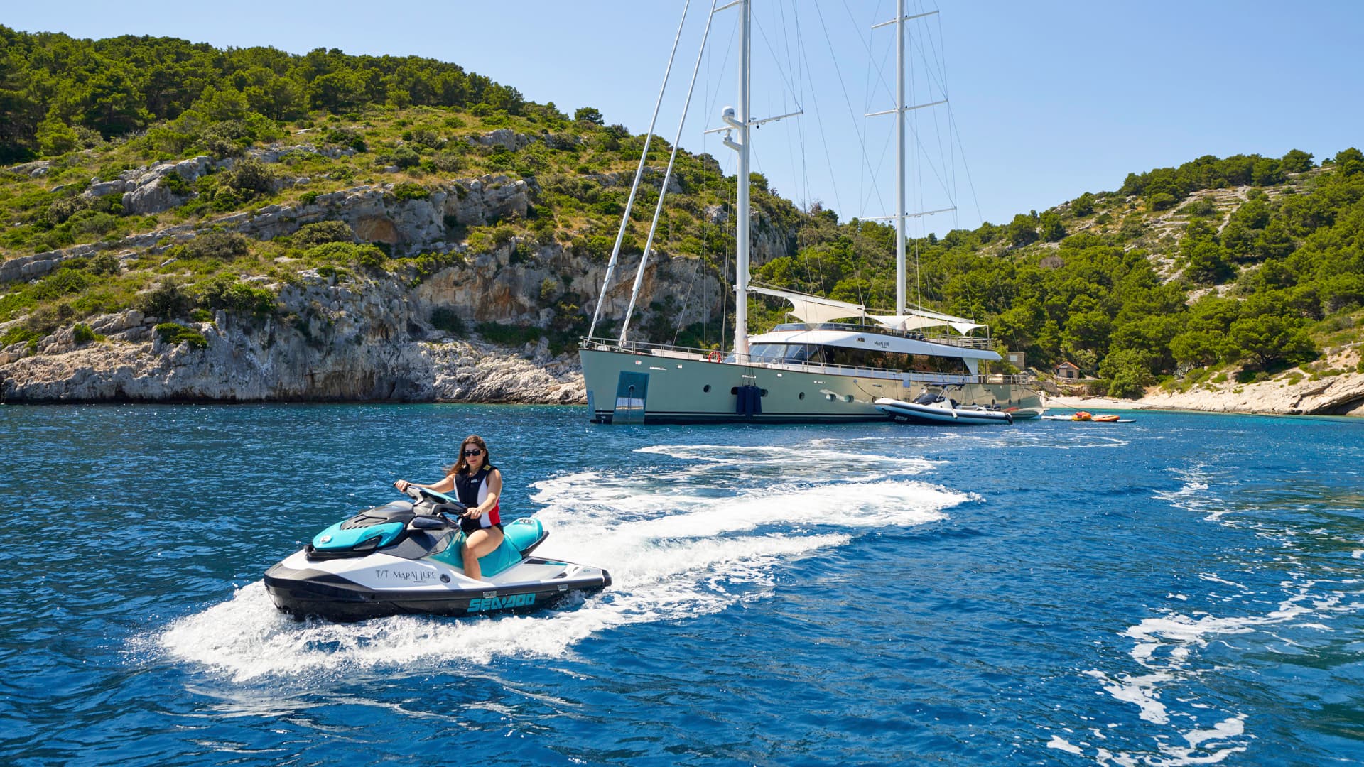 Woman riding a jet ski on blue water near a large sailboat anchored by a rocky, green coastline.