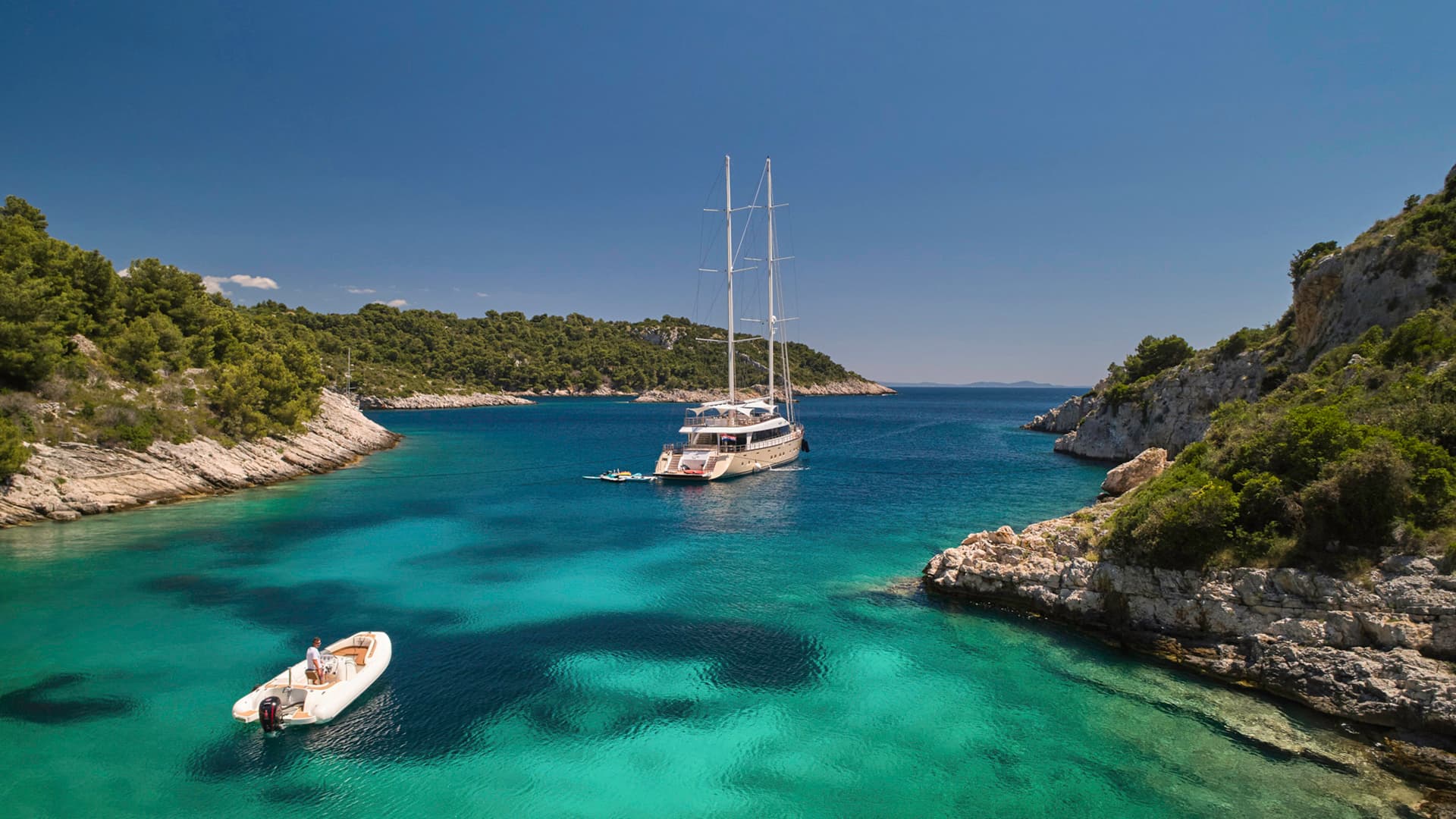 Large sailboat anchored in turquoise cove with rocky, forested shoreline and small dinghy.