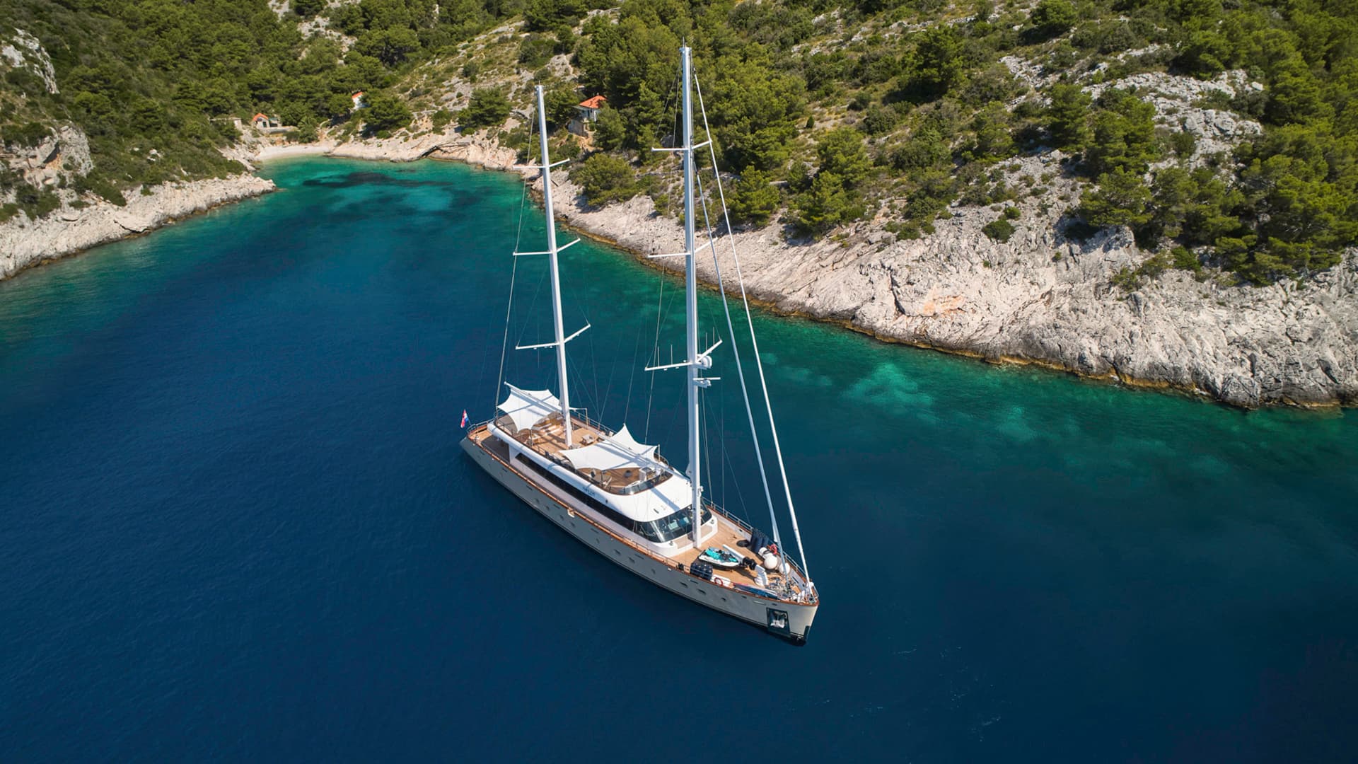 Large sailing yacht anchored in deep blue cove near rocky, forested coastline.