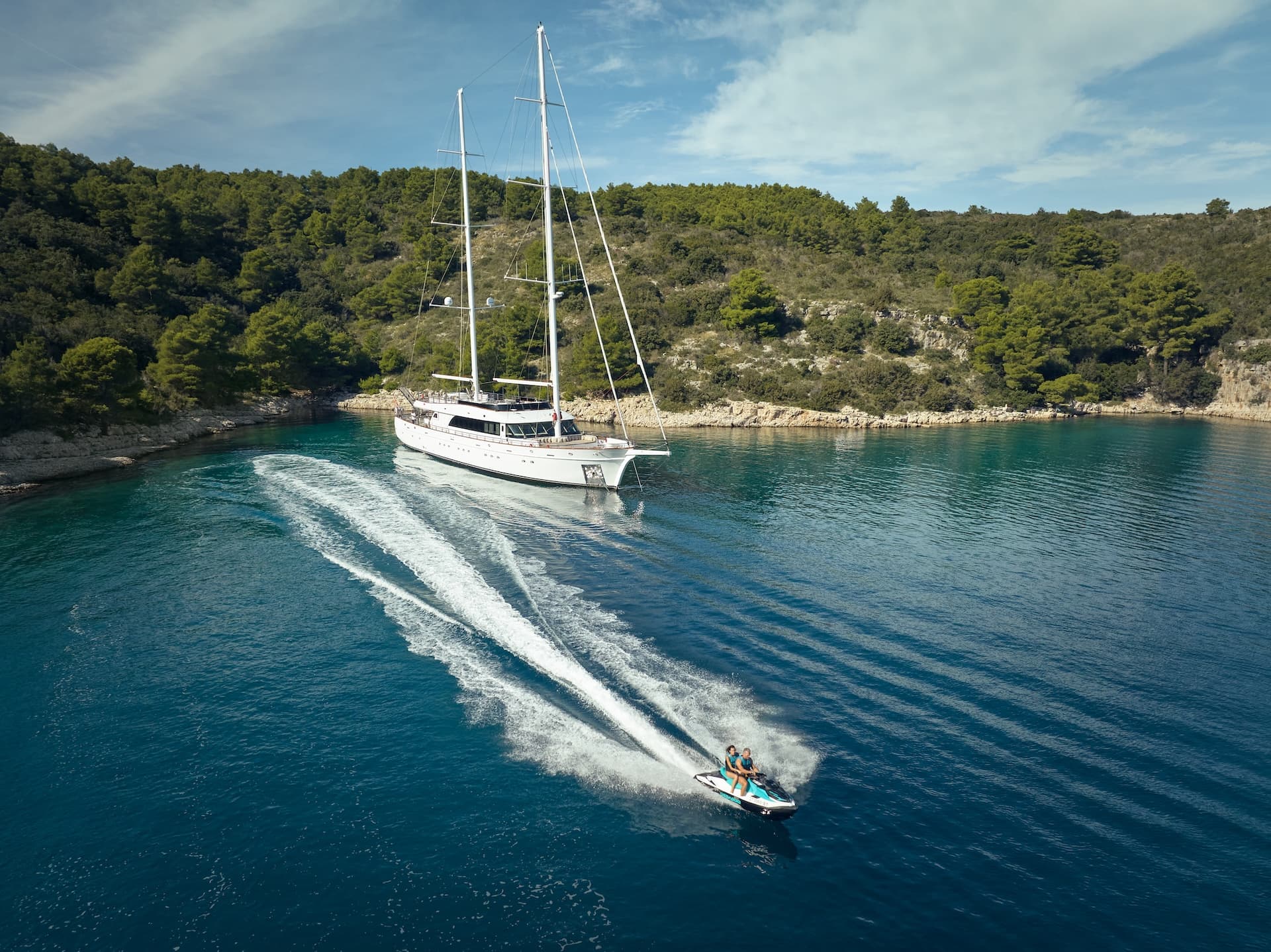People jet skiing near large sailboat anchored by forested coastline.