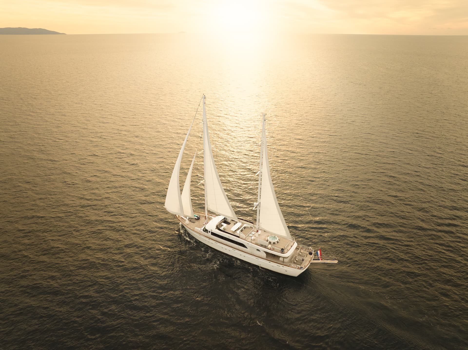 Large white sailing yacht with sails up on dark water at sunset near distant island.