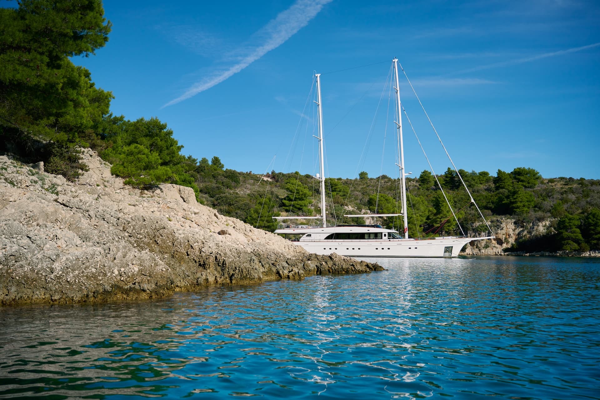 Large white sailboat anchored near rocky, tree-covered coastline under blue sky.