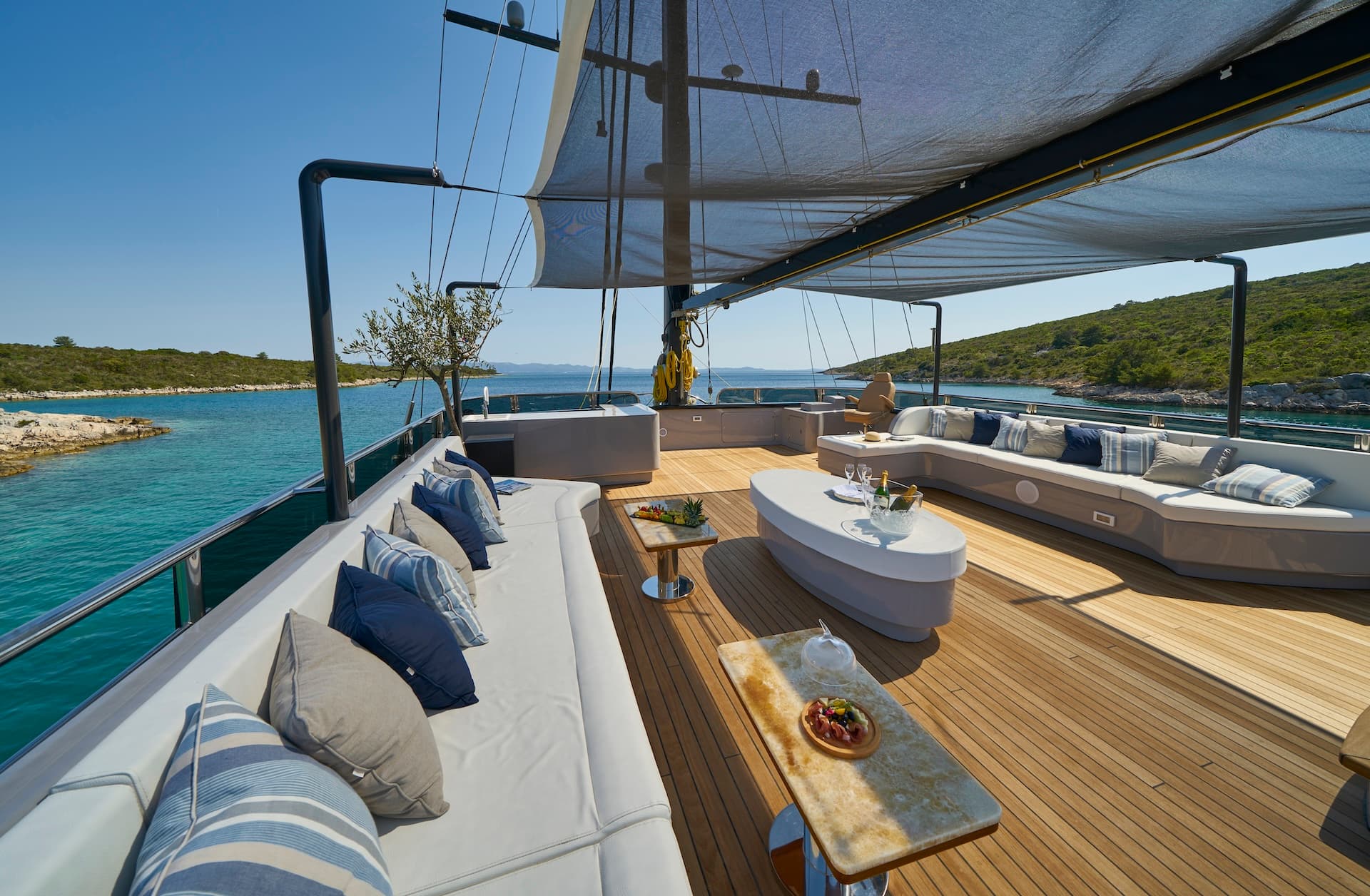 Yacht deck lounge with white seating, teak flooring, and snacks anchored near a green, rocky coastline.