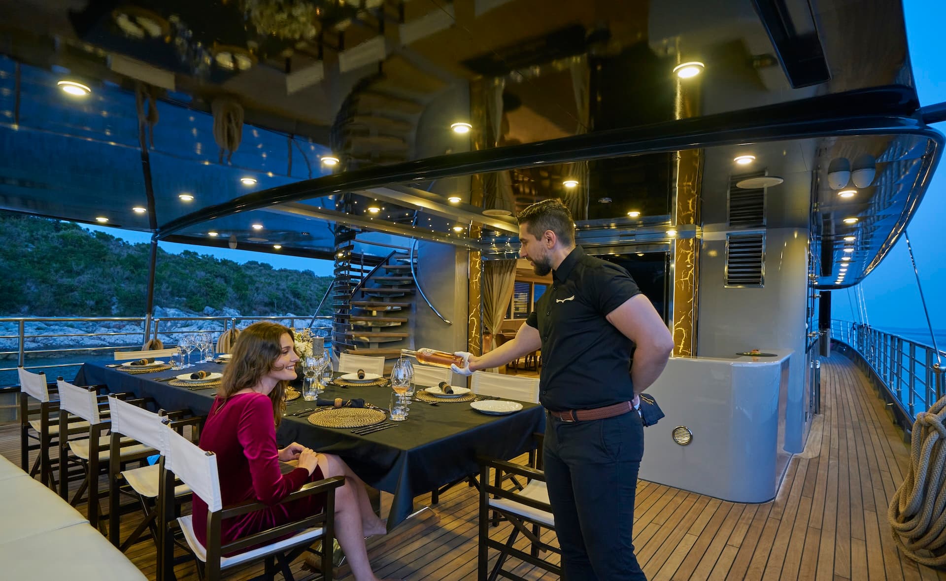 Yacht dining deck with steward serving wine near a wooded coastline at dusk