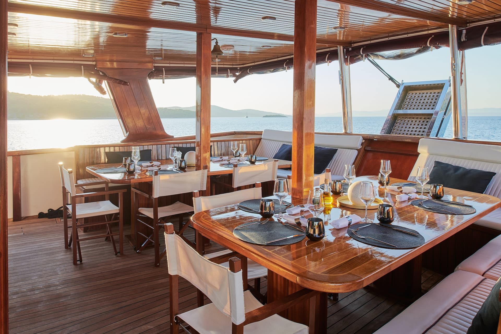 Dining area on a wooden yacht with set tables overlooking the sea at sunset.