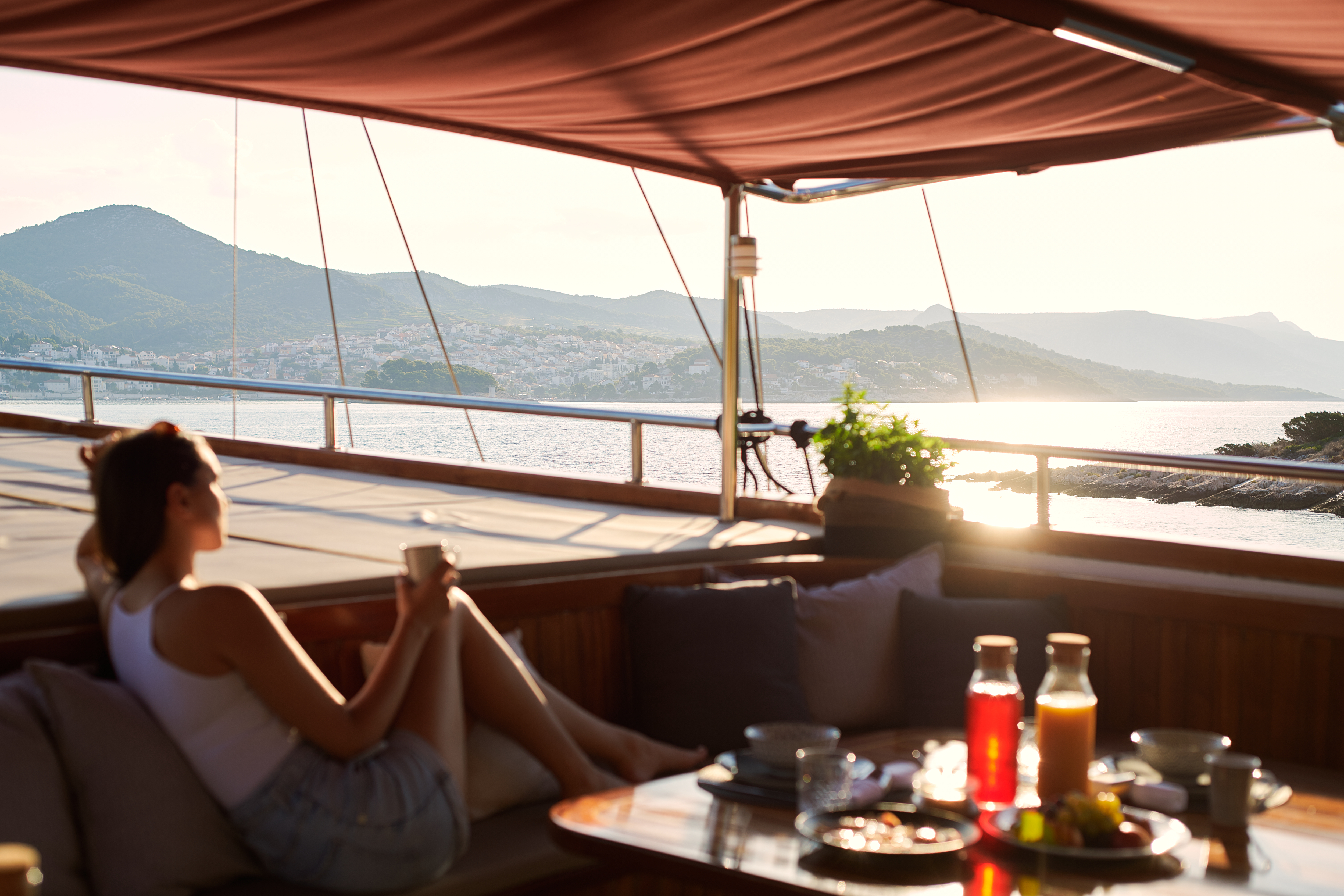 Woman relaxing on yacht deck with breakfast overlooking coastal town and mountains.