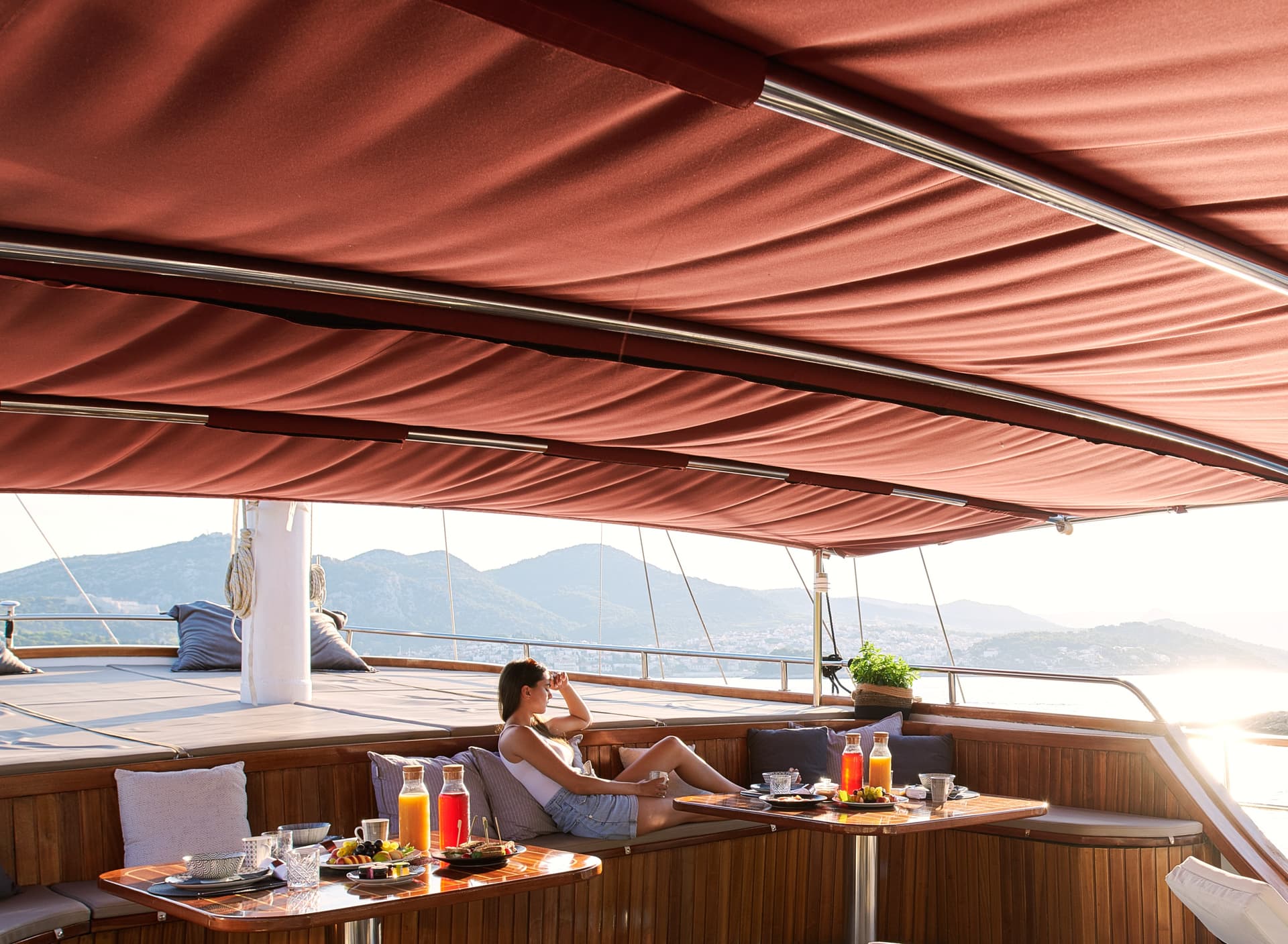 Woman relaxing on yacht deck with breakfast, overlooking mountains and sunlit coastline.
