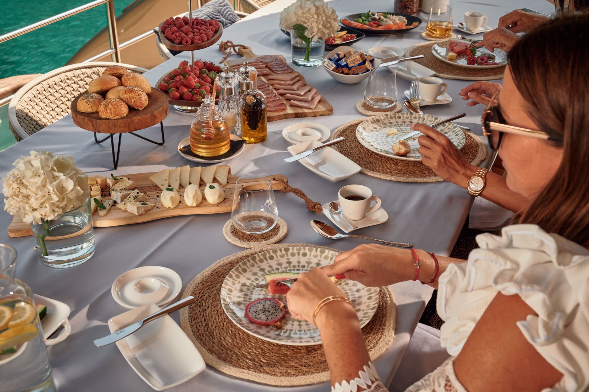People dining on a yacht with fresh fruit, cheese, cold cuts, and coffee overlooking turquoise water.