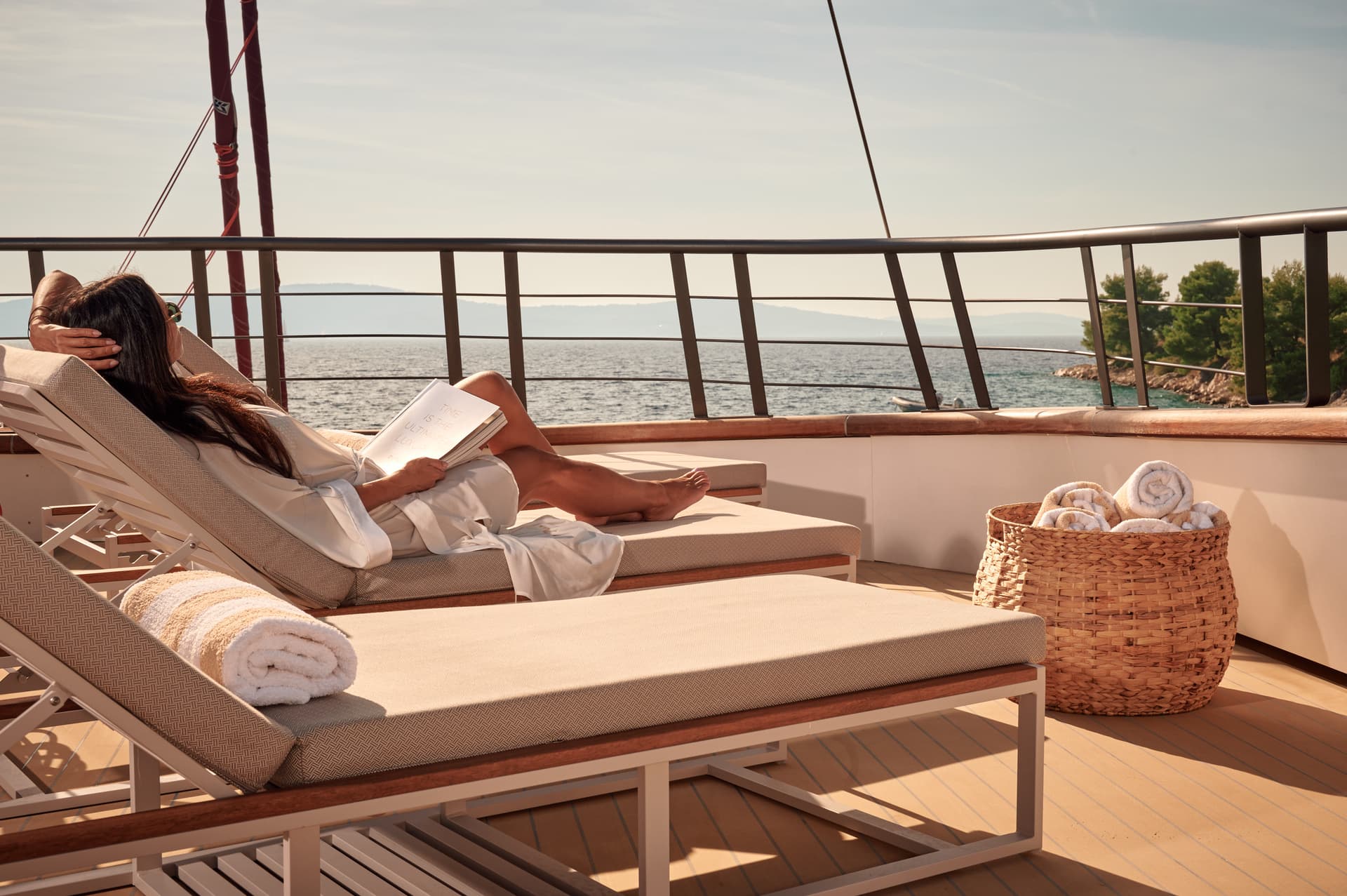 Woman reading on deck chair on yacht overlooking calm sea and distant coastline